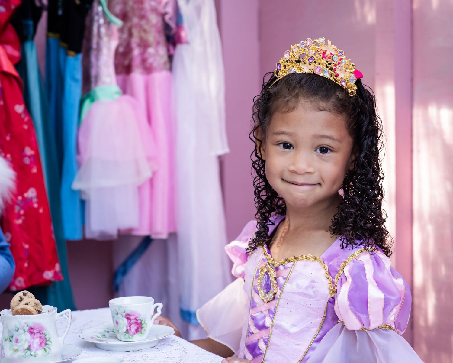 Children in princess dresses and tiaras pose happily during dress-up time at a pink-themed party celebration.