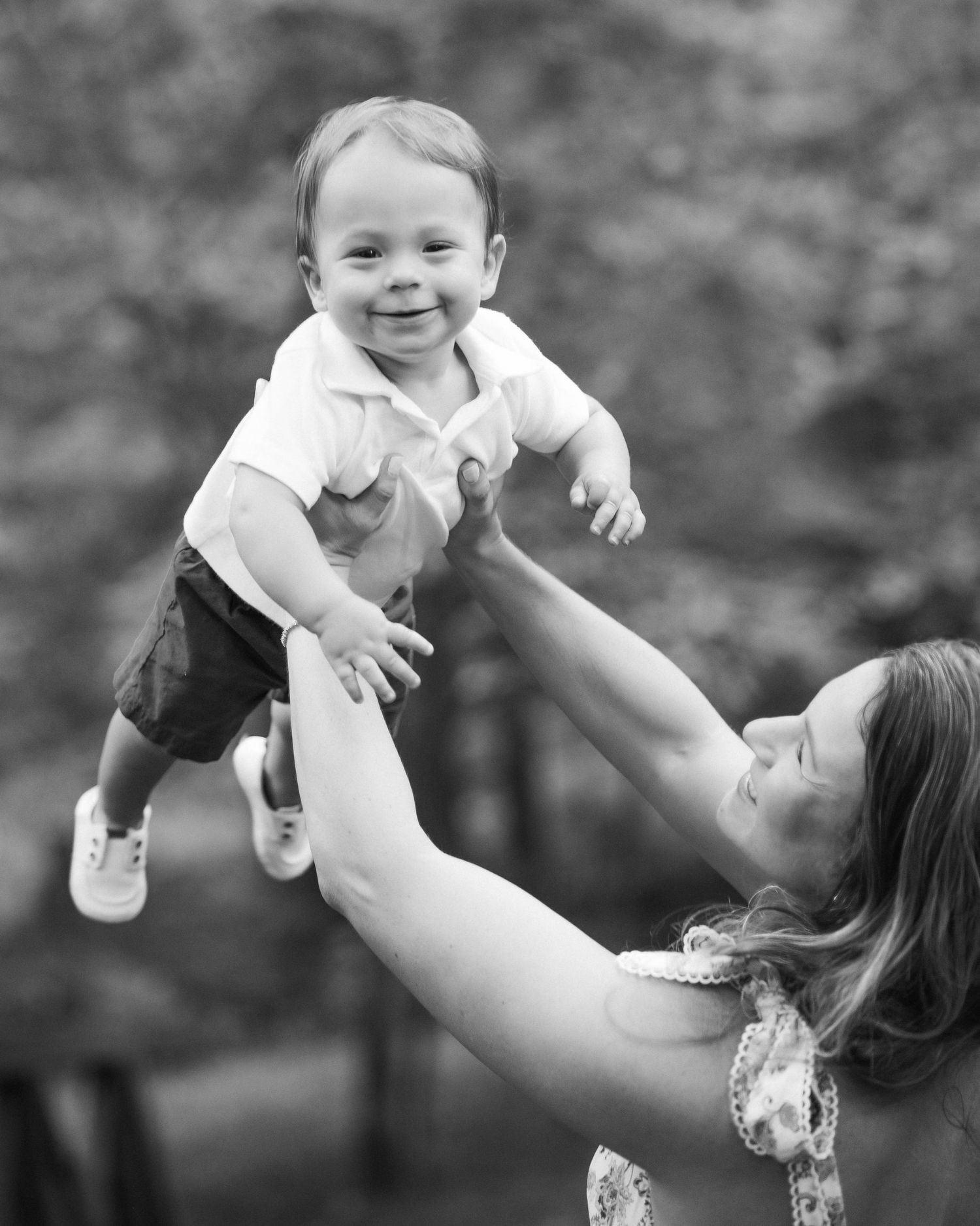 Black and white photo of a smiling baby being lifted in the air by a woman outdoors.