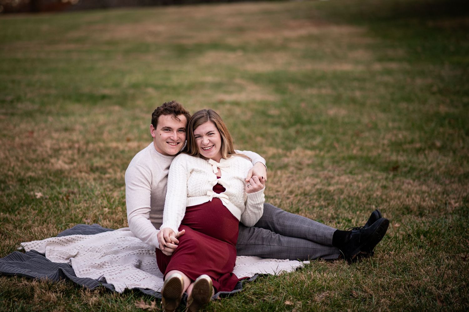 Couple sitting on blanket in grass, woman in red skirt and white jacket, man in white shirt, outdoor maternity photo.