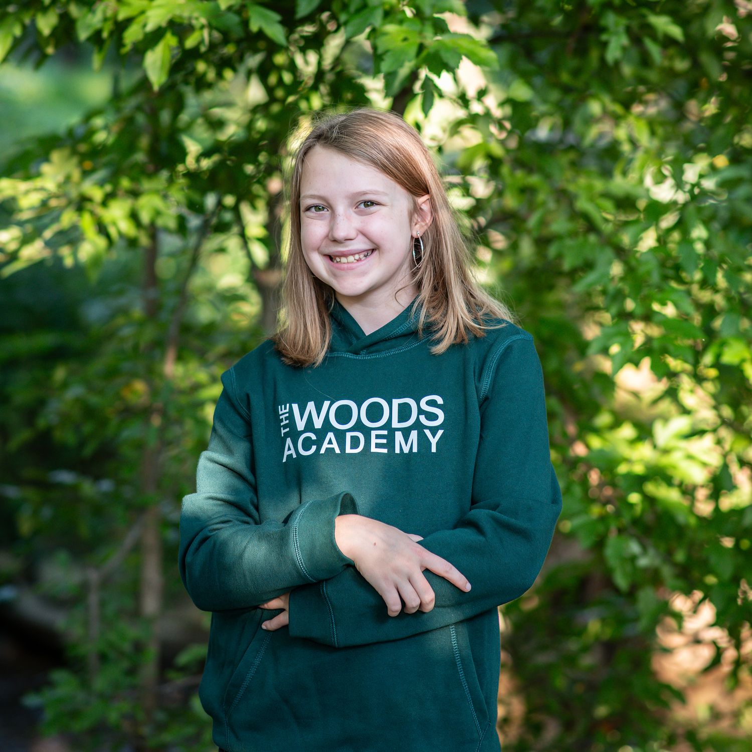 Smiling student wearing a green Woods Academy hoodie poses outdoors surrounded by lush green trees.
