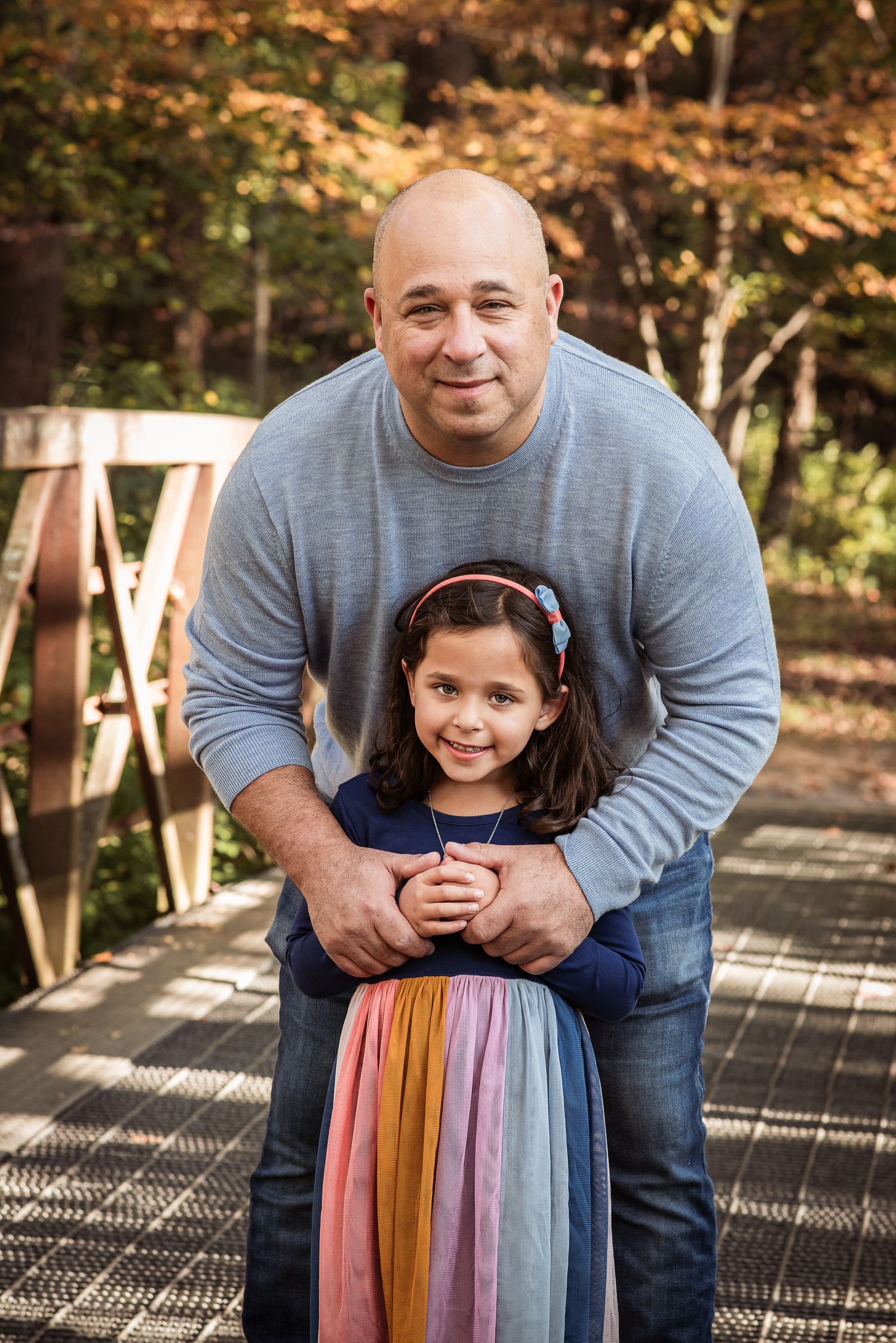 A bald man smiles warmly while hugging a young girl in a colorful skirt on a wooden bridge outdoors.