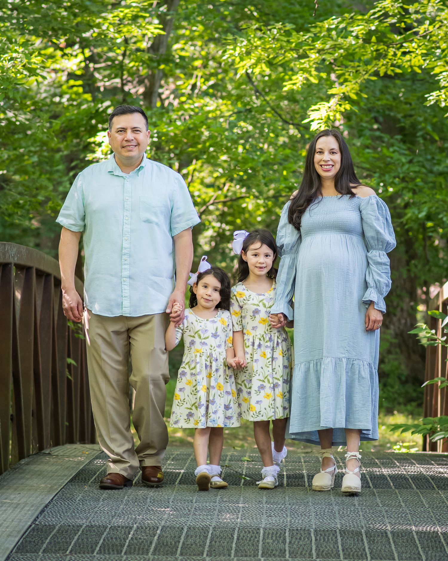 Family of four posing on a bridge surrounded by lush green trees, parents and two young girls in matching floral dresses.