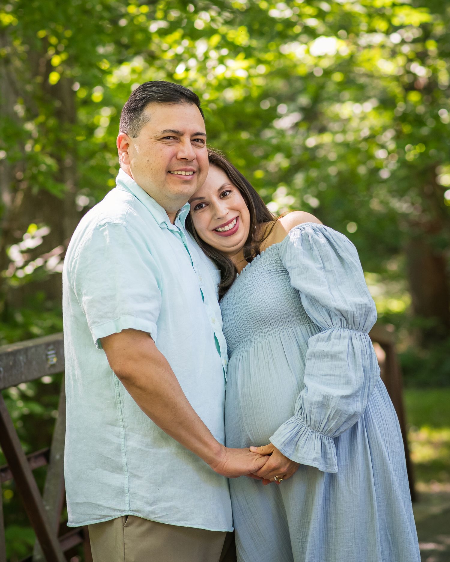 Smiling couple posing outdoors in green park setting, pregnant woman in light blue dress held by man in white shirt.