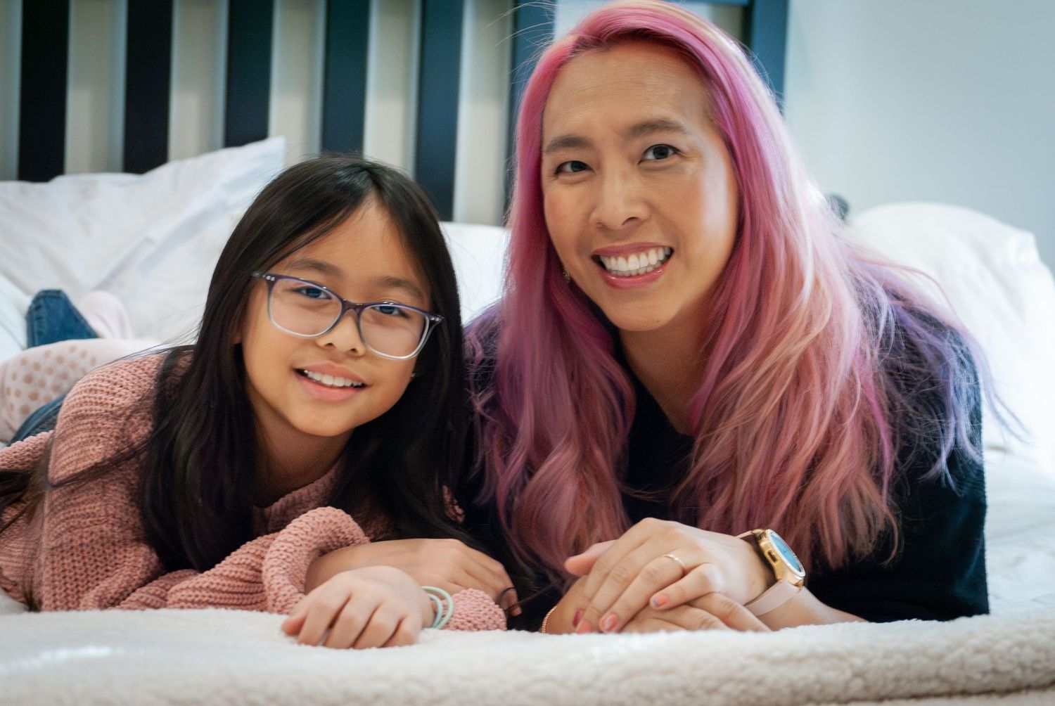 Two people lying on a bed sharing a happy moment together, one with vibrant pink hair.