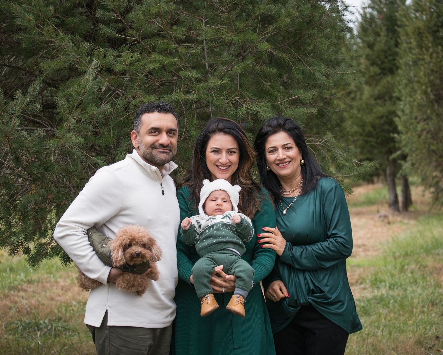 Family of three adults and a baby posing outdoors with a fluffy dog among pine trees in a forest setting.