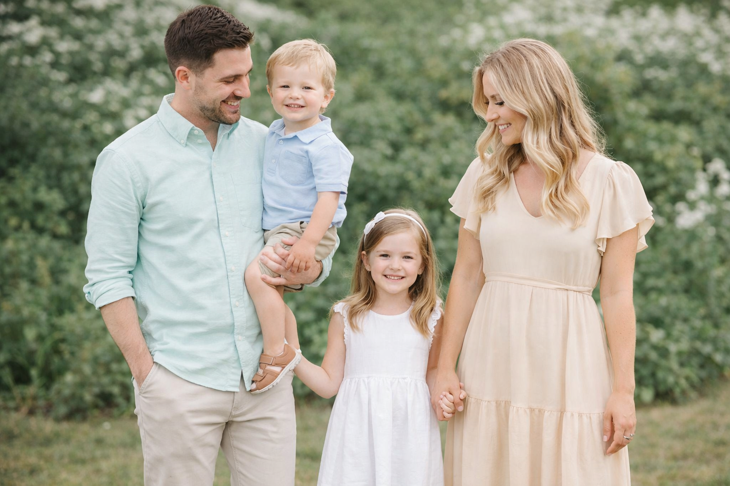 Family wearing coordinated pastel outfits poses together in a lush green outdoor setting during springtime.