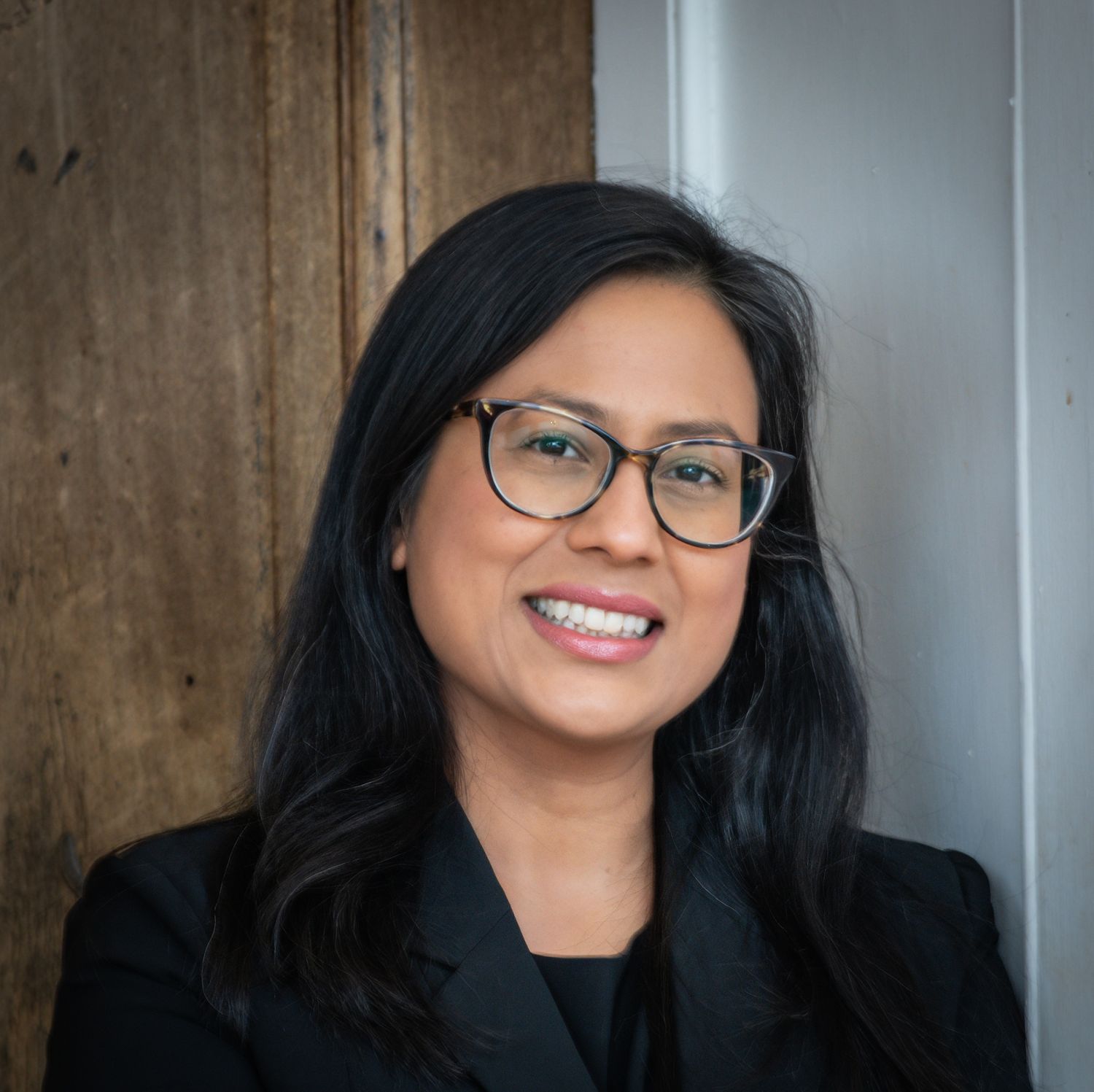 Professional headshot of someone with long dark hair and glasses smiling warmly against a gray background.