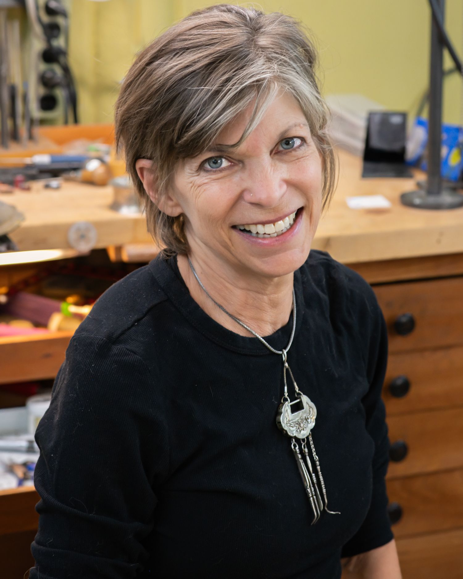 Smiling woman with short gray hair wearing a black top and silver necklace poses in a jewelry workshop.