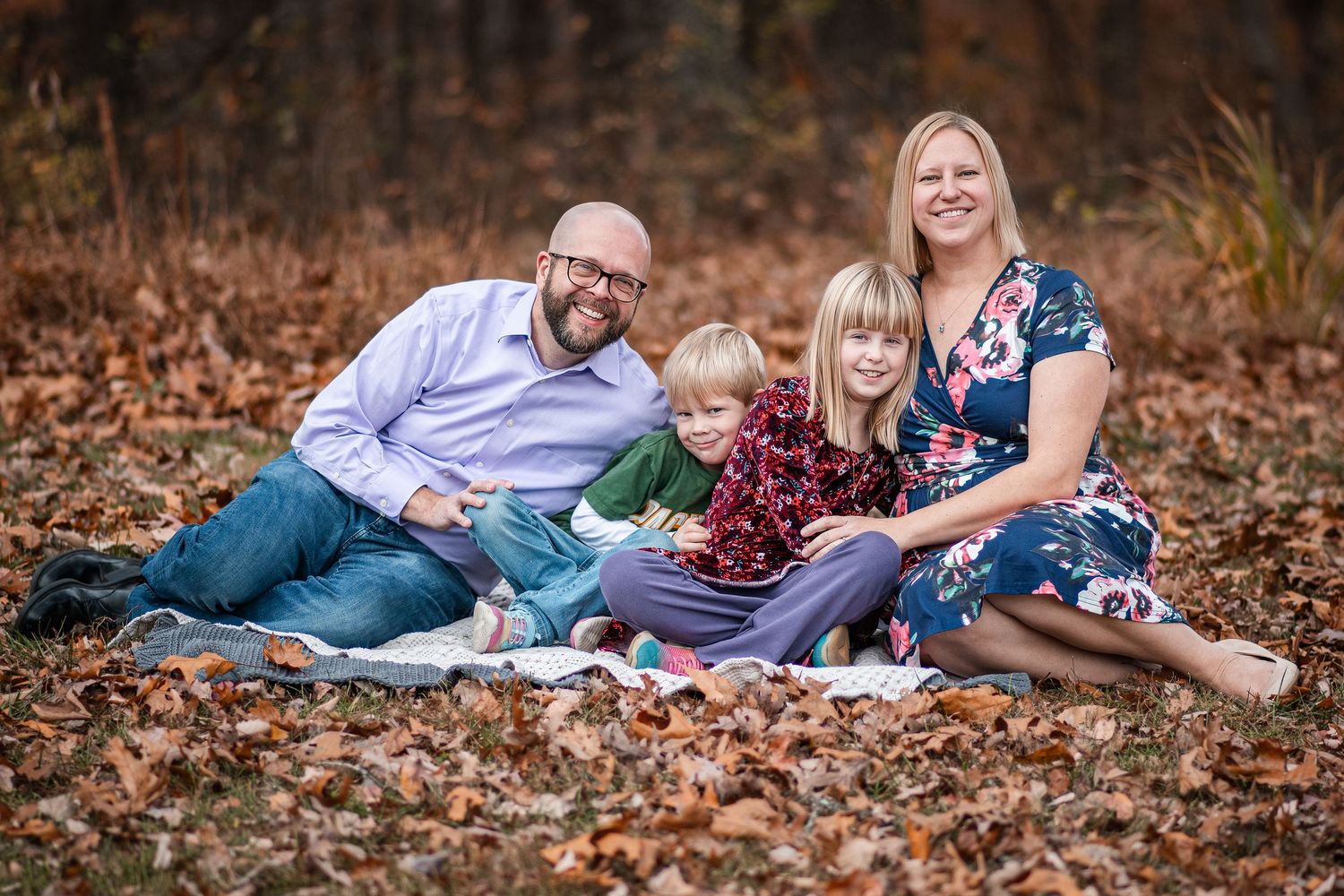 Family of four smiling together on a blanket surrounded by autumn leaves in a wooded outdoor setting.