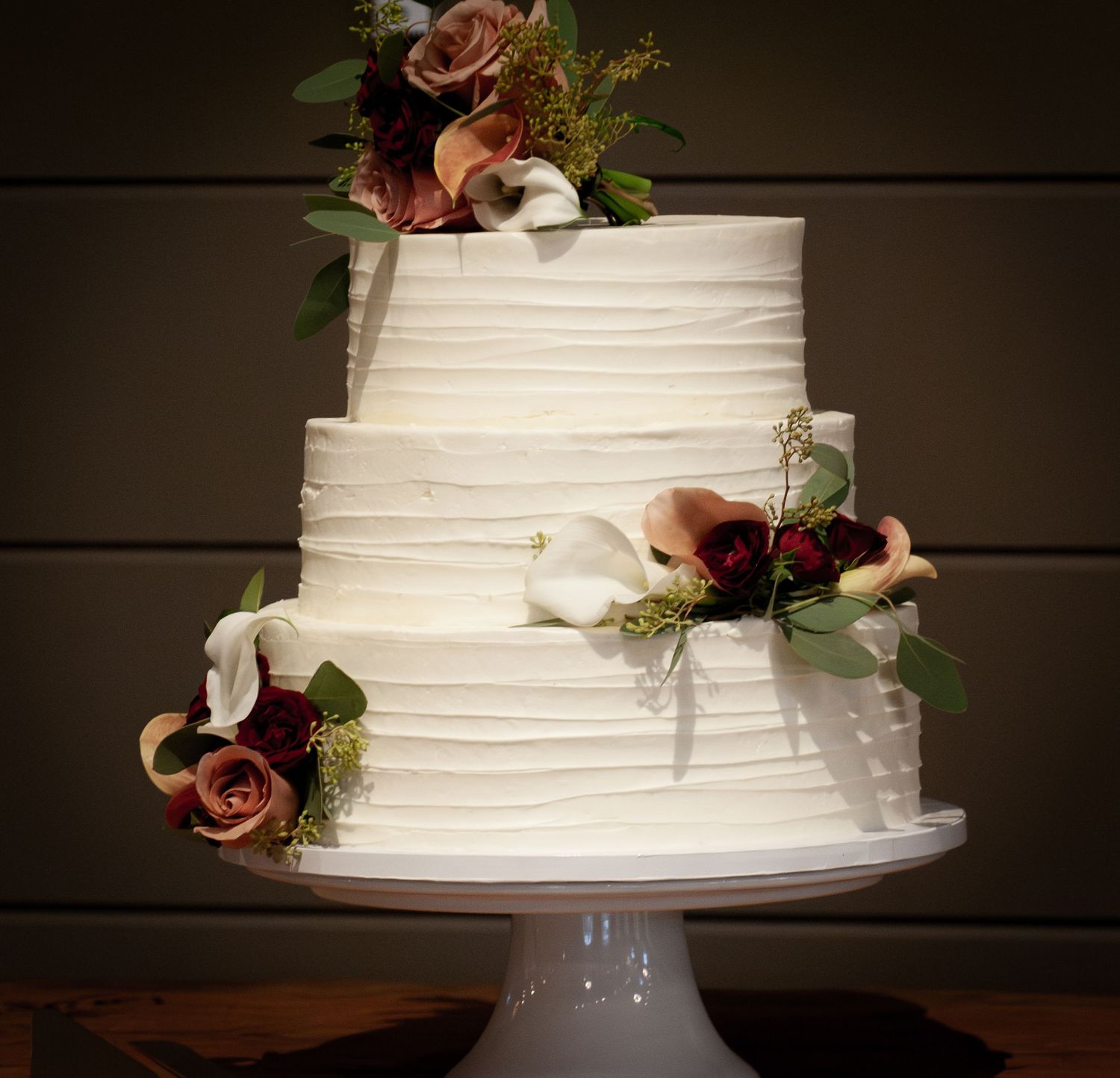 White textured wedding cake with mauve roses and greenery displayed on a white pedestal against a dark background.