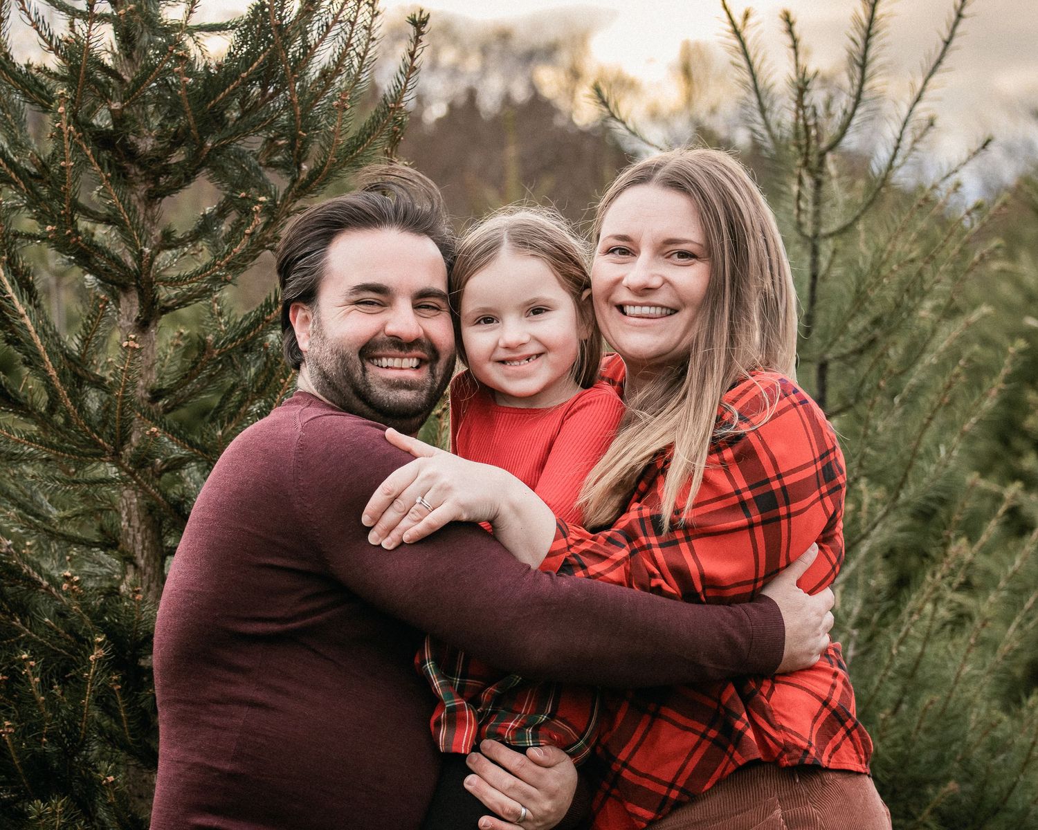 A family wearing burgundy and red plaid outfits poses together among evergreen Christmas trees at sunset.
