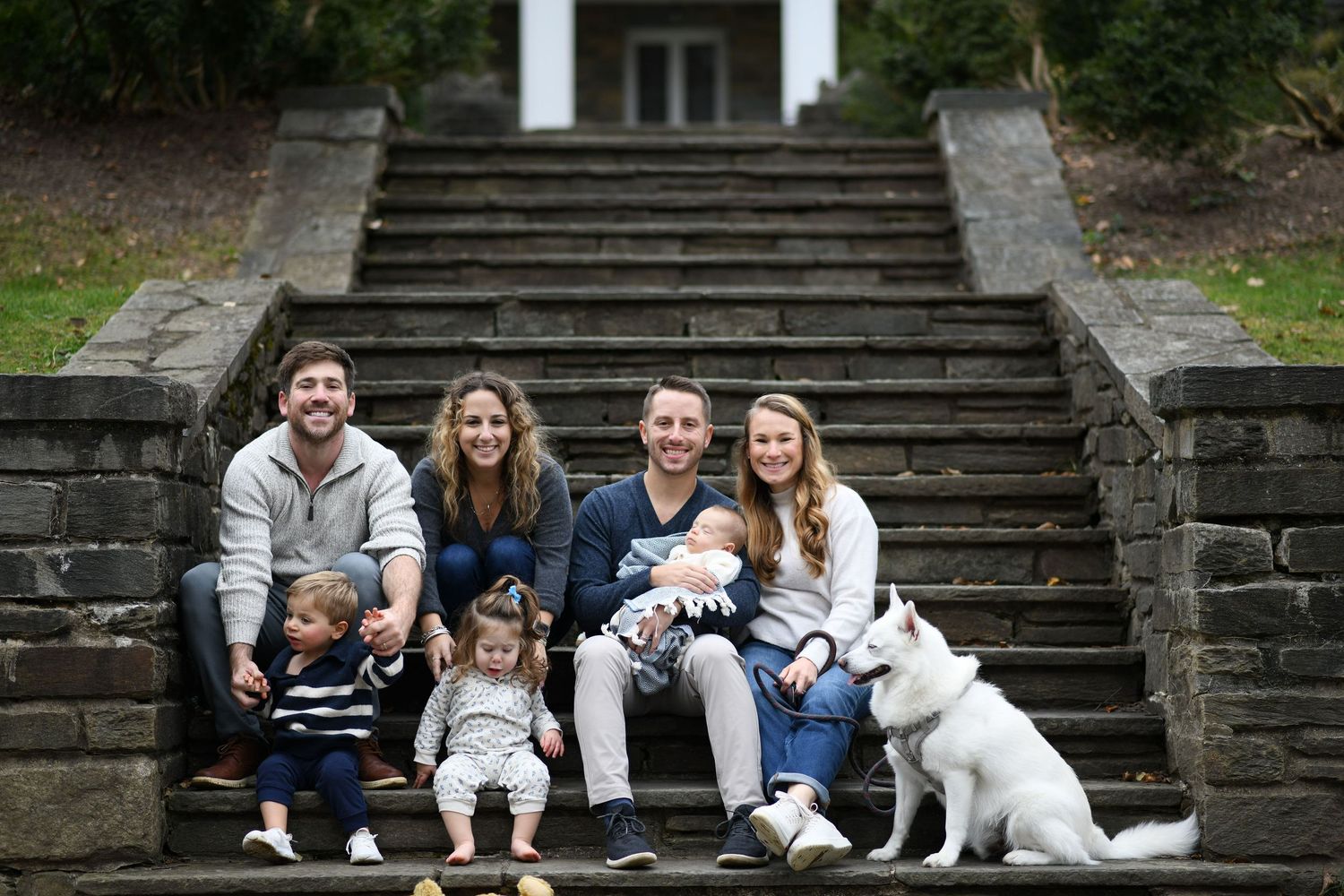 A family sits on stone steps outdoors with their white dog for a portrait session.