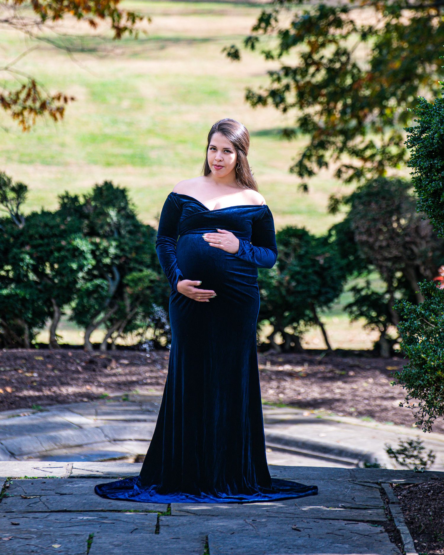 Pregnant woman in elegant navy blue velvet off-shoulder gown posing outdoors in a lush green garden setting.