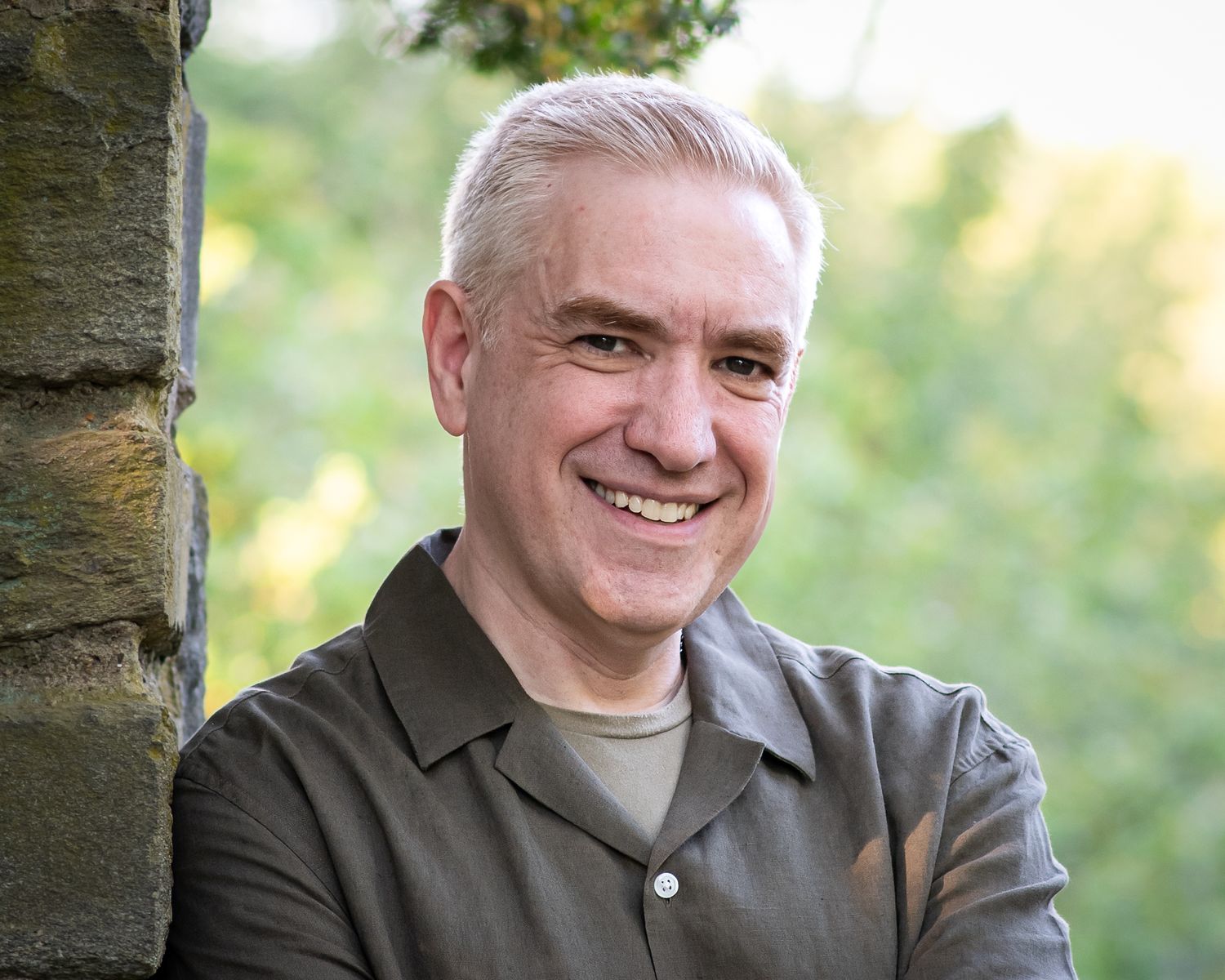 Smiling middle-aged man with gray hair leaning against a stone pillar outdoors with green trees in background.