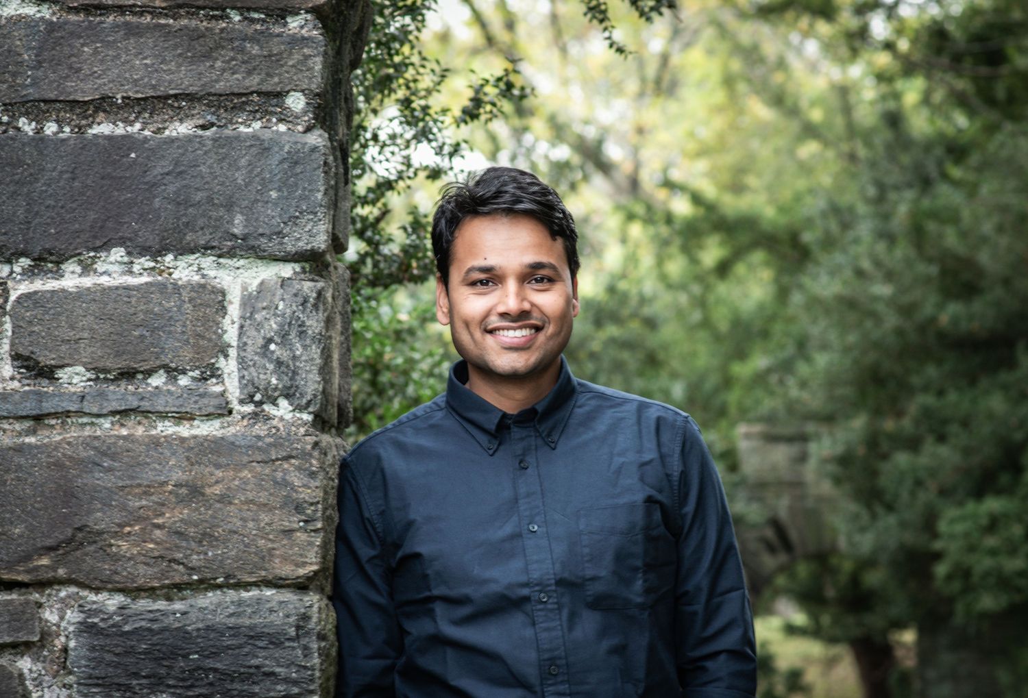 Professional outdoor portrait of a person in a navy blue button-down shirt standing next to a stone wall.