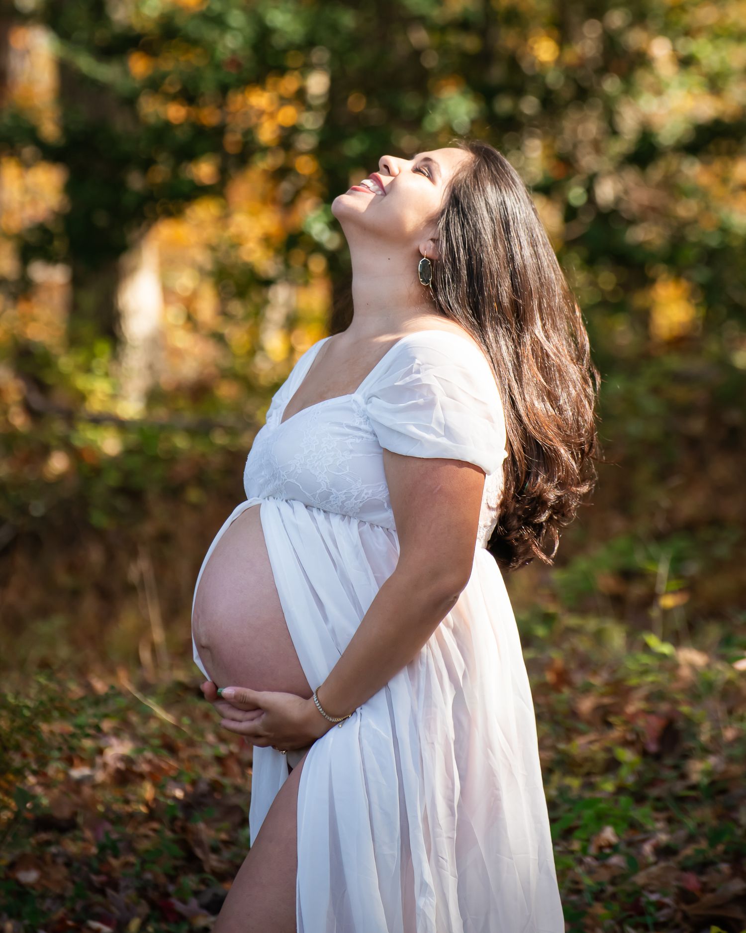 A pregnant woman in a flowing white dress poses in a sunlit autumn forest setting.
