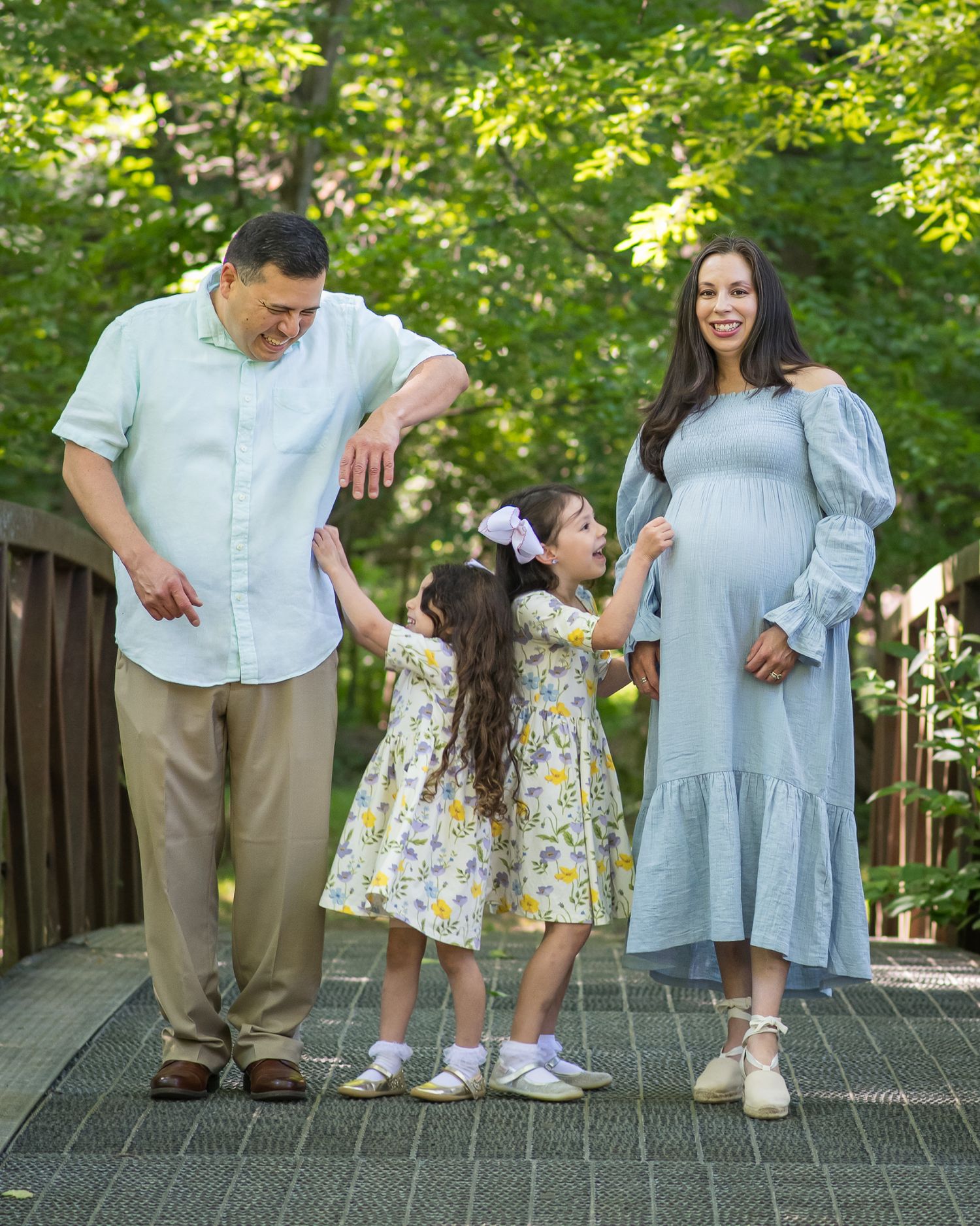 Happy family maternity photo on a bridge, parents holding toddler girl's hands outdoors in lush green park.