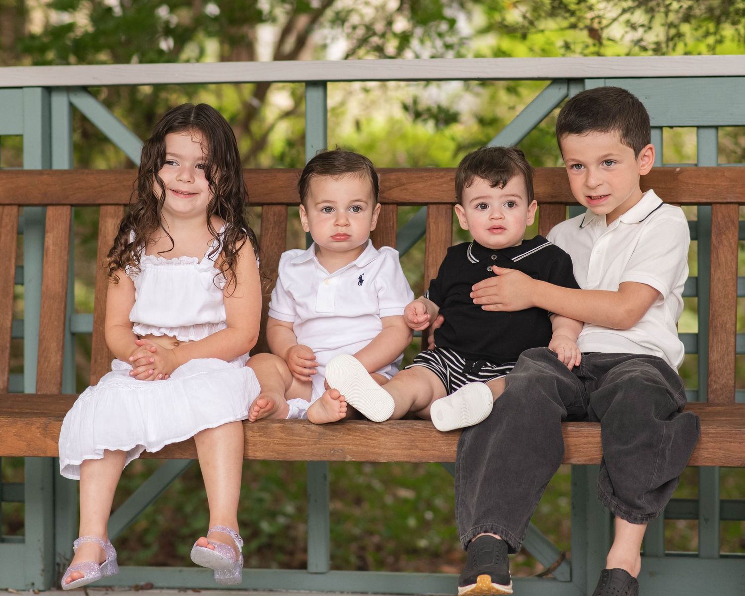 Four young children sitting together on a wooden bench outdoors, smiling for a family portrait photo.