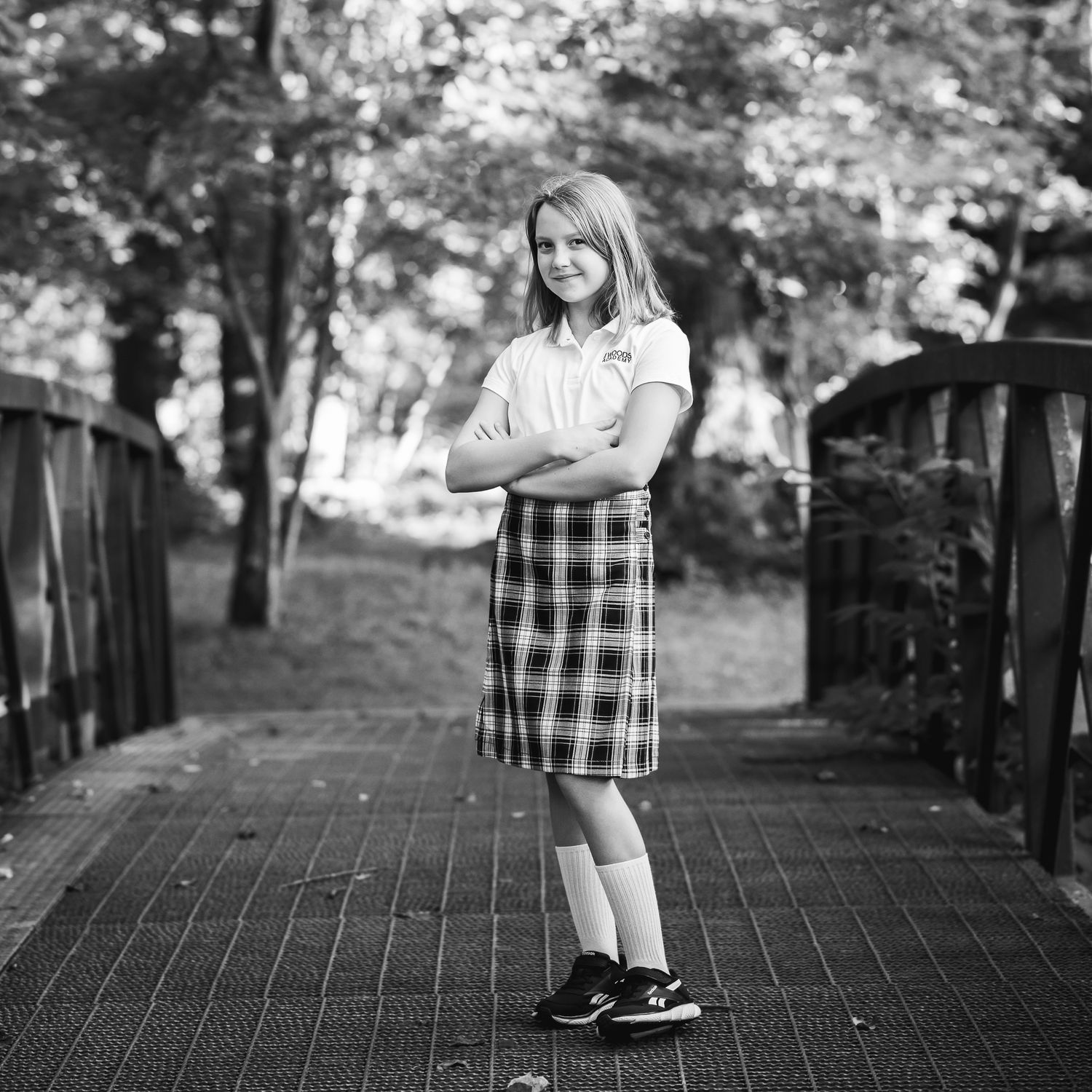 Black and white photo of a young girl in plaid skirt and white shirt standing on a wooden bridge.