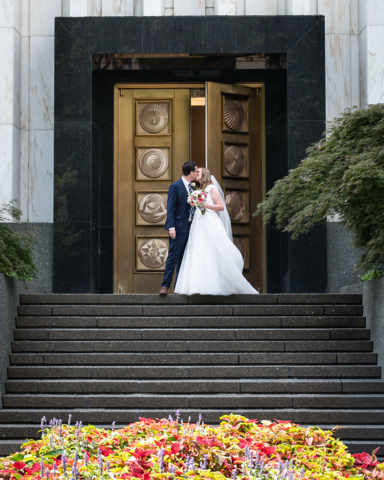 Bride and groom kissing on steps of elegant building with ornate gold doors and colorful flowers in foreground.