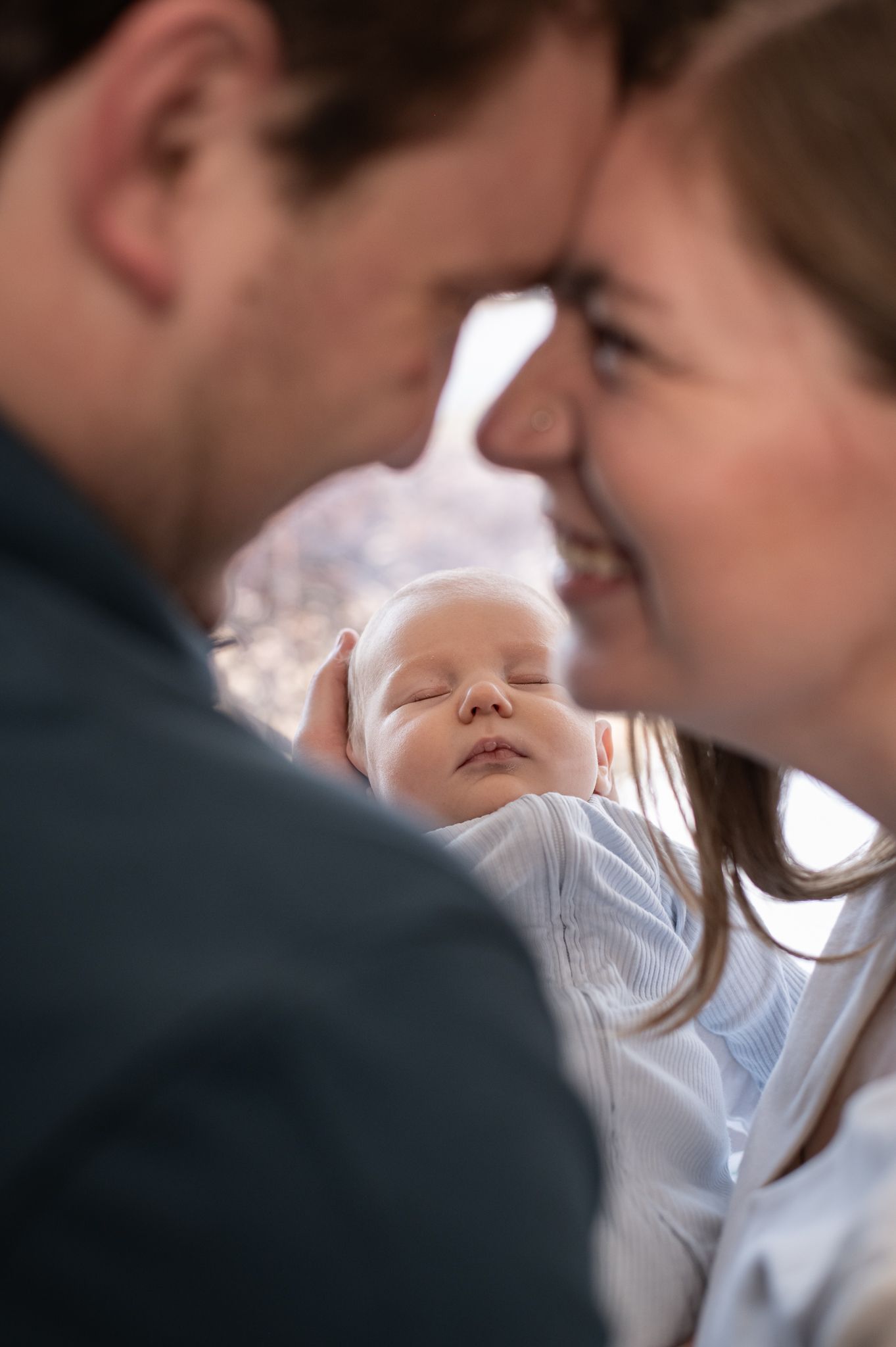 Parents tenderly touch foreheads while cradling their sleeping newborn baby wrapped in a white swaddle.
