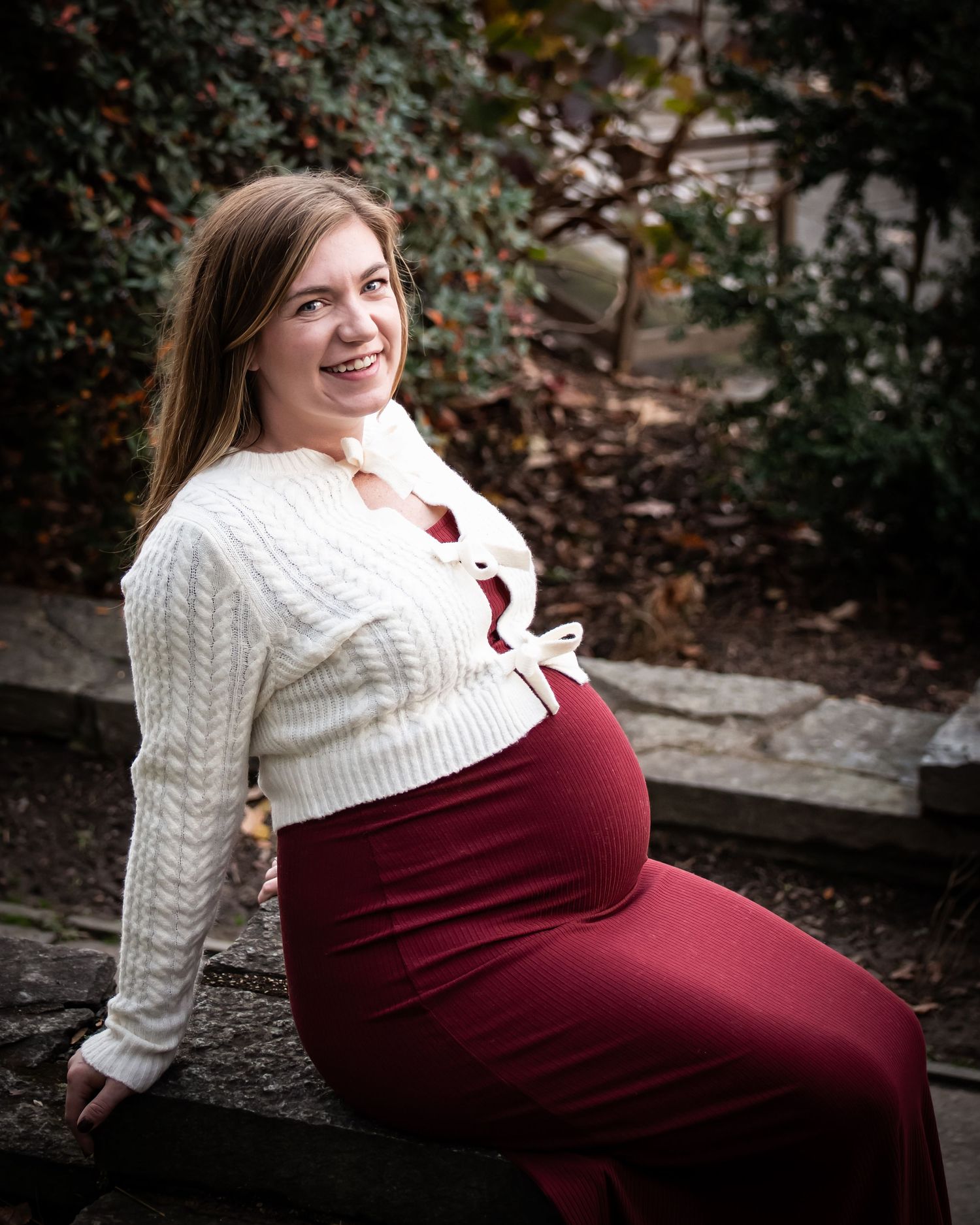 Maternity photo session in a garden setting with someone wearing a burgundy dress and cream knit sweater.