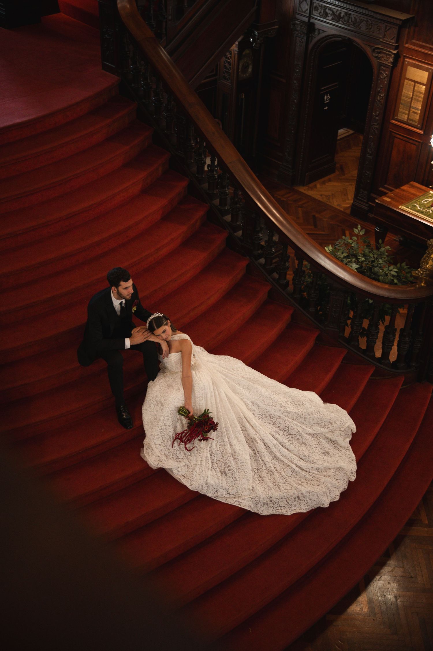Bride and groom pose on grand red carpeted staircase, her lace ball gown trailing elegantly in a luxurious venue.