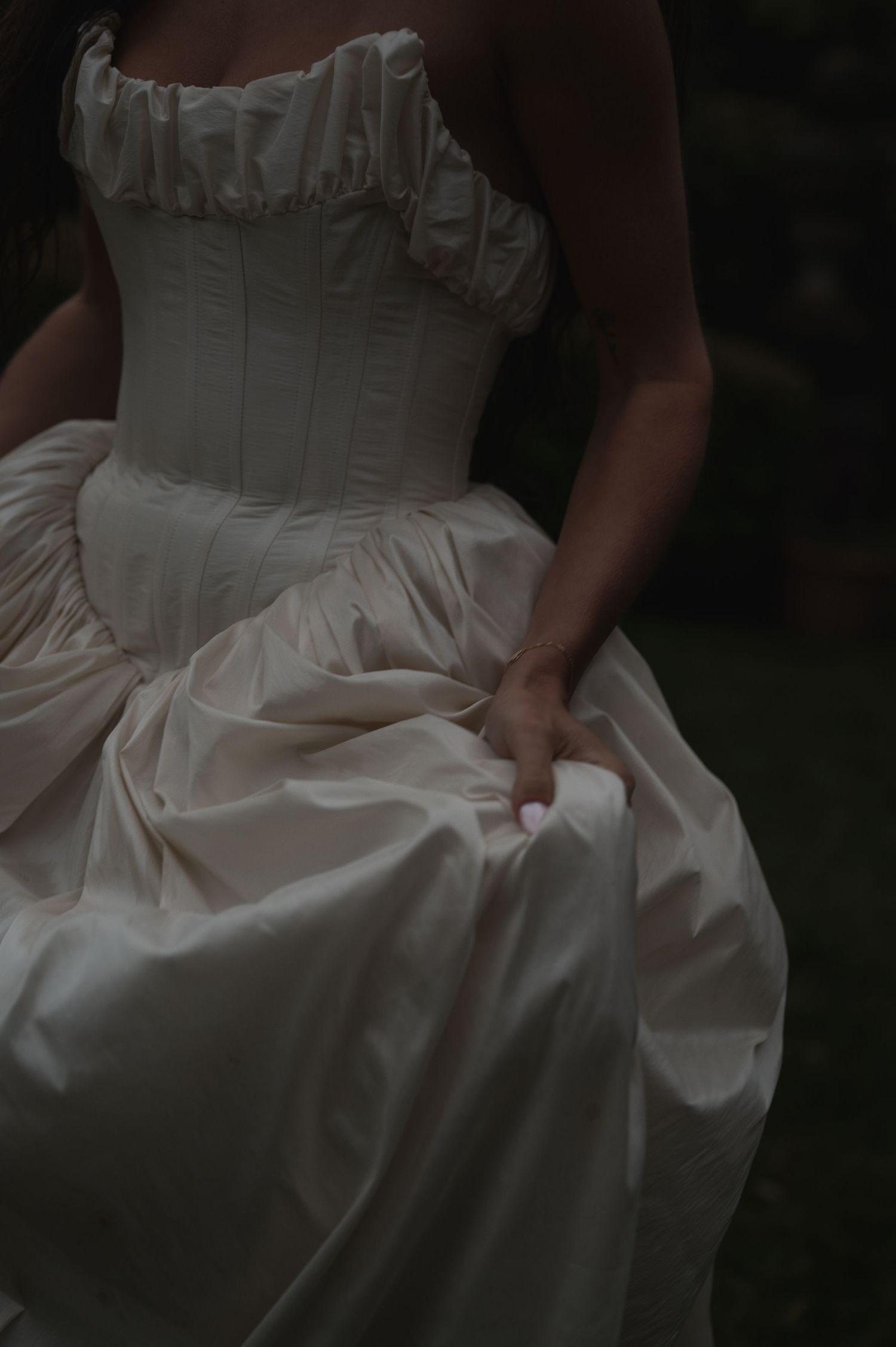 A woman in an elegant ivory satin ballgown corset dress gathers her full skirt with her hand in a moody dark setting.