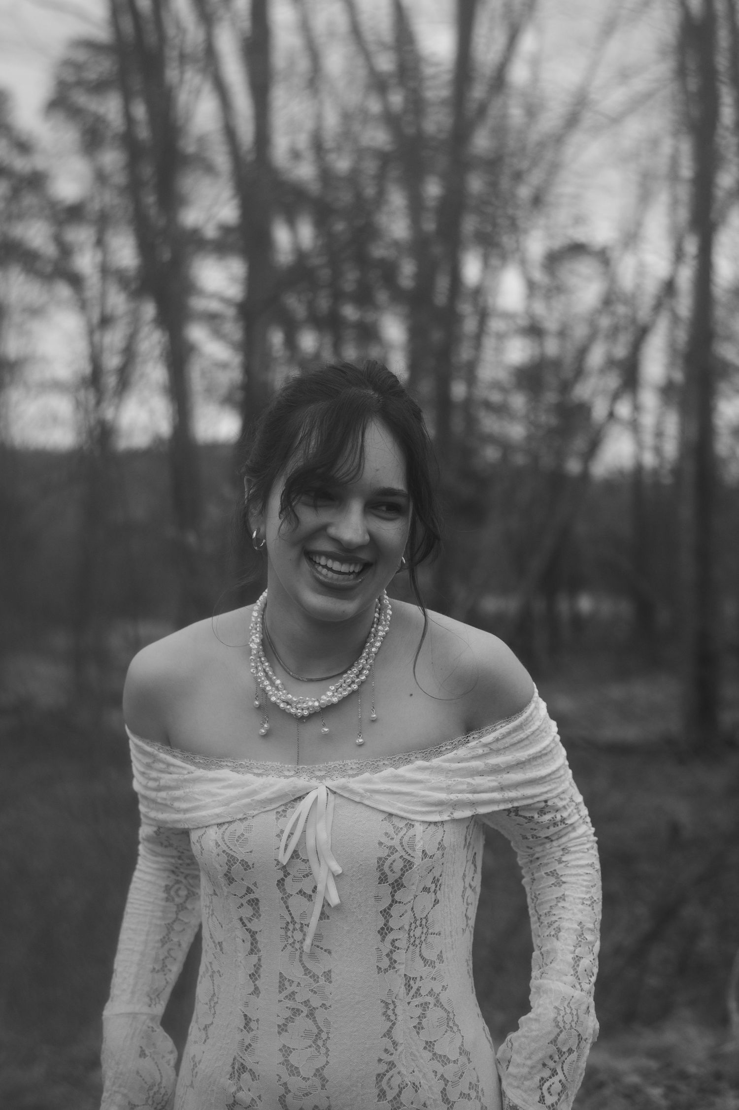Smiling bride in off-shoulder lace wedding dress and pearl necklace poses outdoors among bare trees in black and white photo.