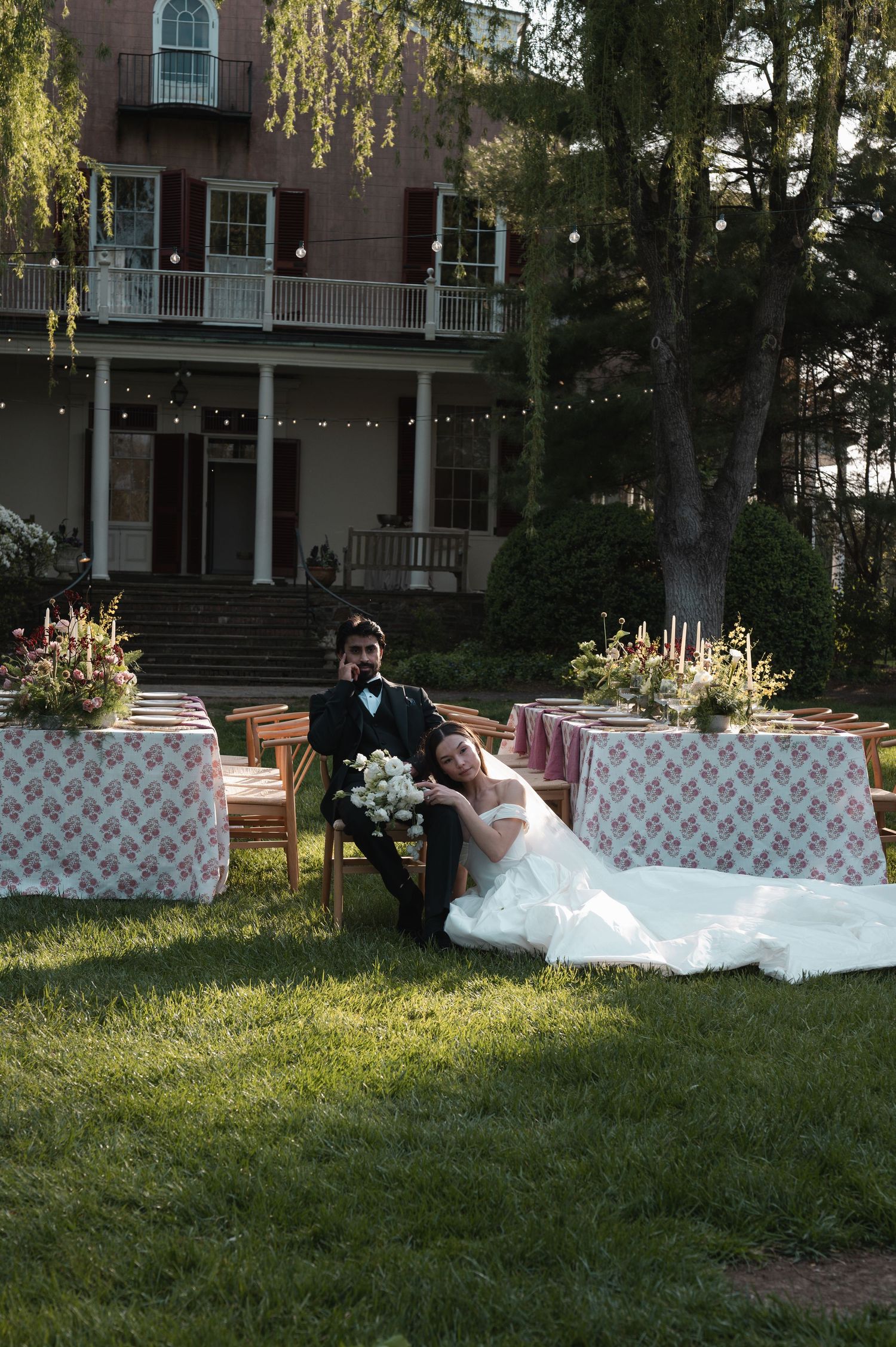 Bride and groom sitting together on lawn at outdoor wedding reception with pink floral tablecloths and string lights.