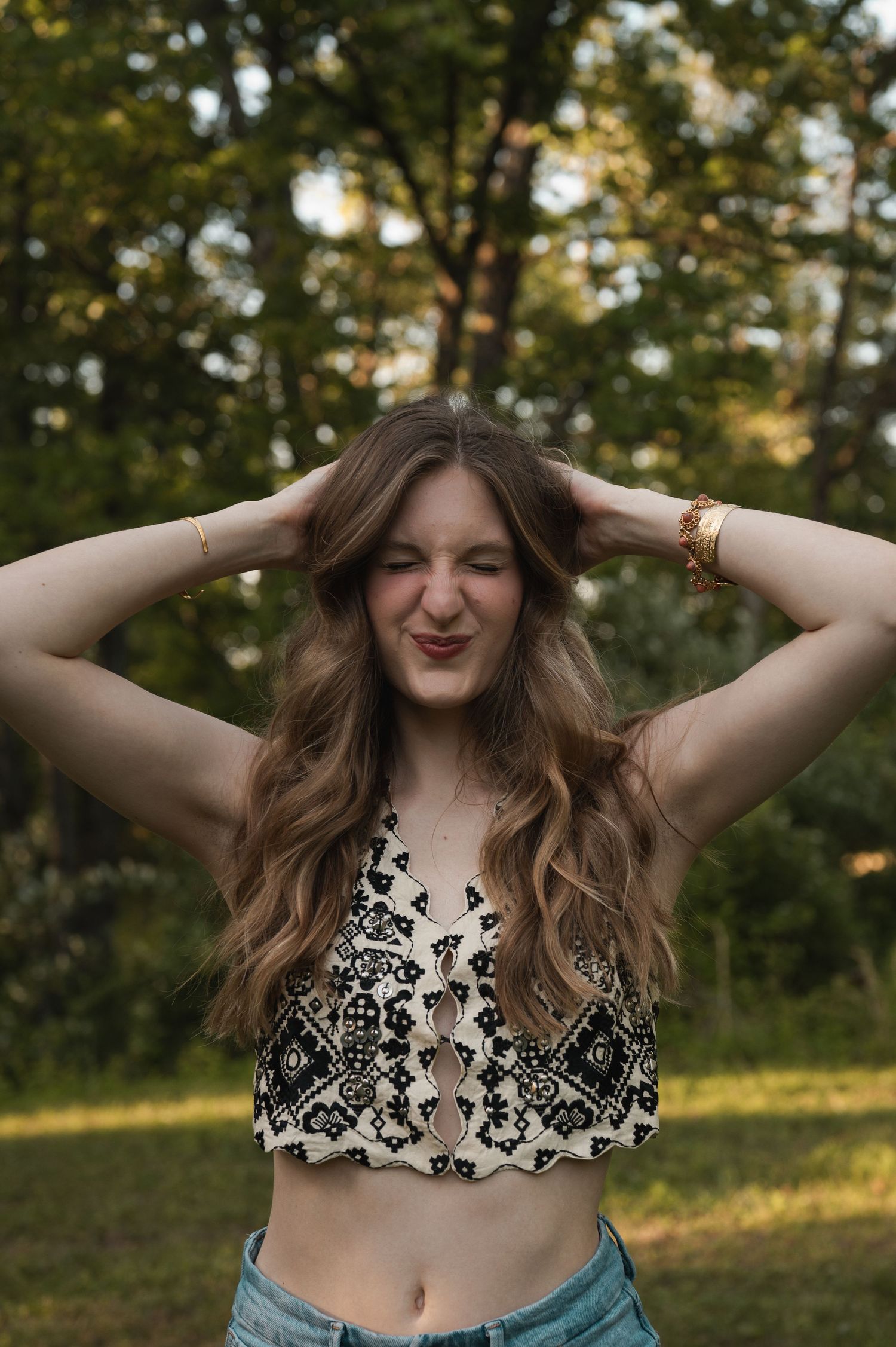 Young woman in crop top posing outdoors with hands in hair, smiling playfully against green trees.