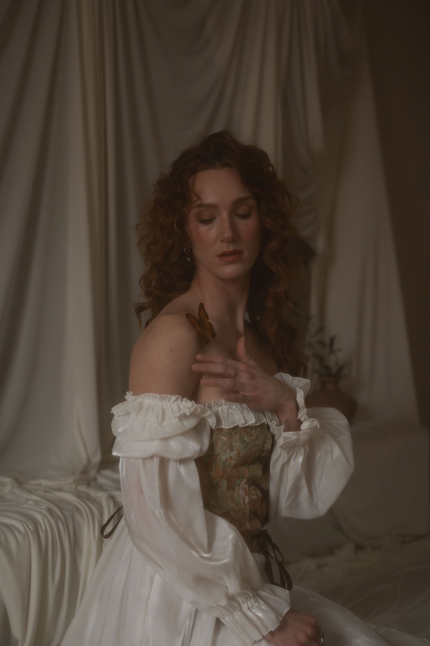 A woman in a white off-shoulder Renaissance gown with corset sits gracefully against draped fabric backdrop.