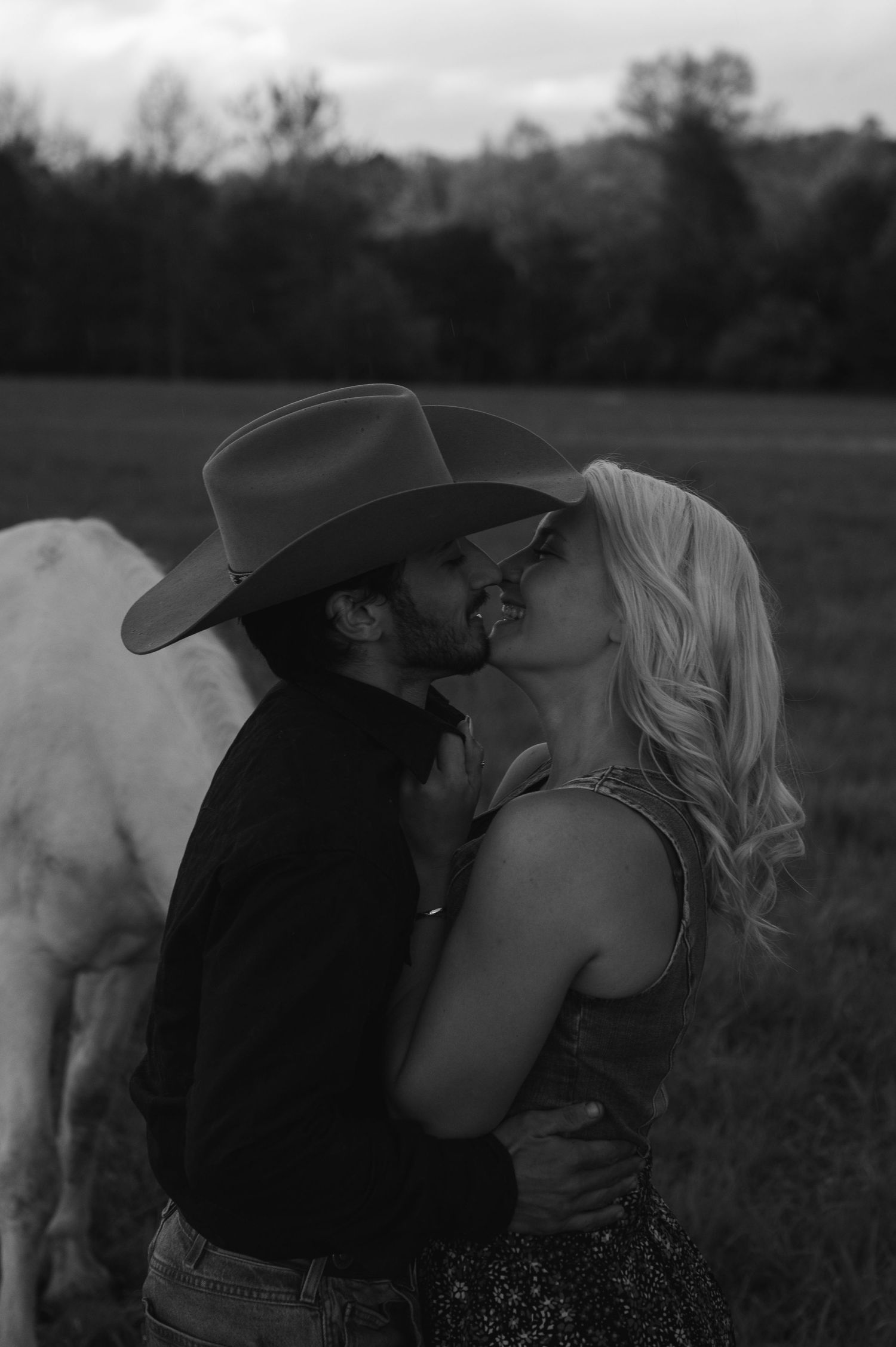 Black and white photo of a couple kissing in a field, man wearing cowboy hat, white horse visible behind them.
