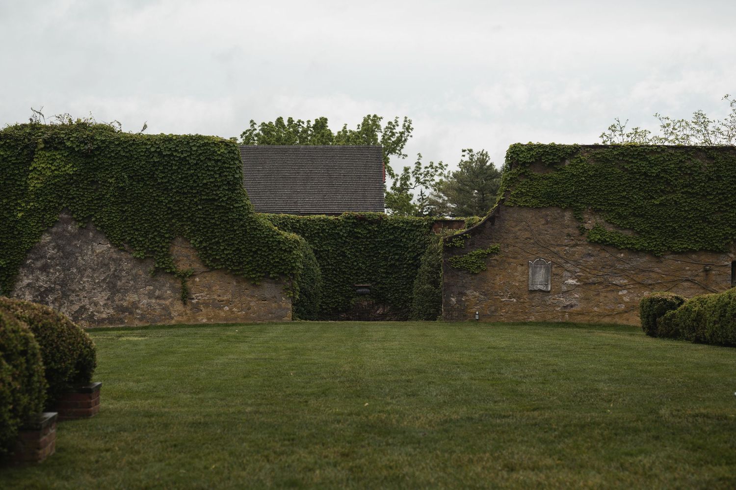 Ivy-covered brick walls surround a lush green lawn with trimmed hedges and a glimpse of a building roof beyond.