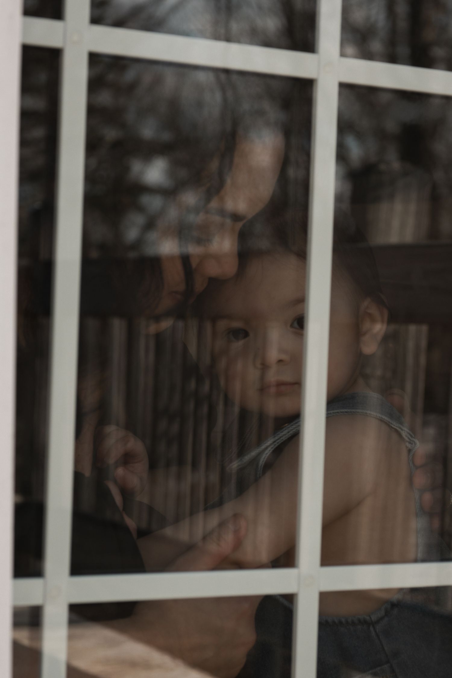 Adult holding a baby while looking through a white-framed window, with trees reflected in the glass.