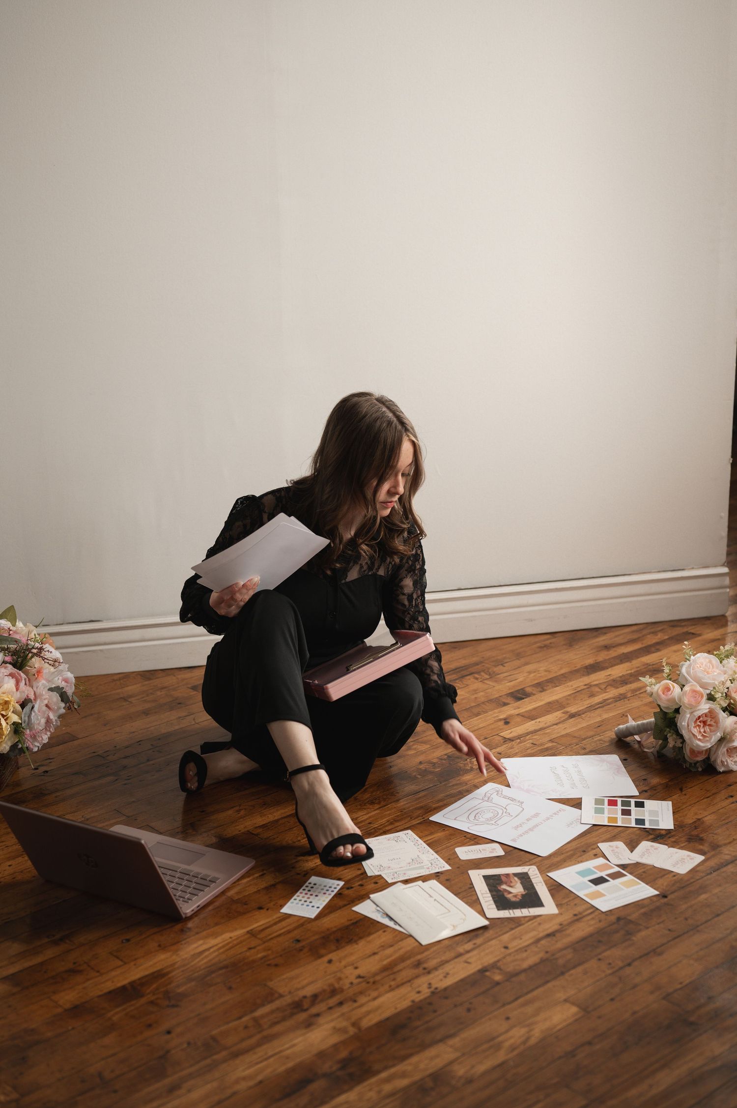 Woman in black outfit sitting on hardwood floor reviewing wedding planning documents beside laptop and floral arrangements.