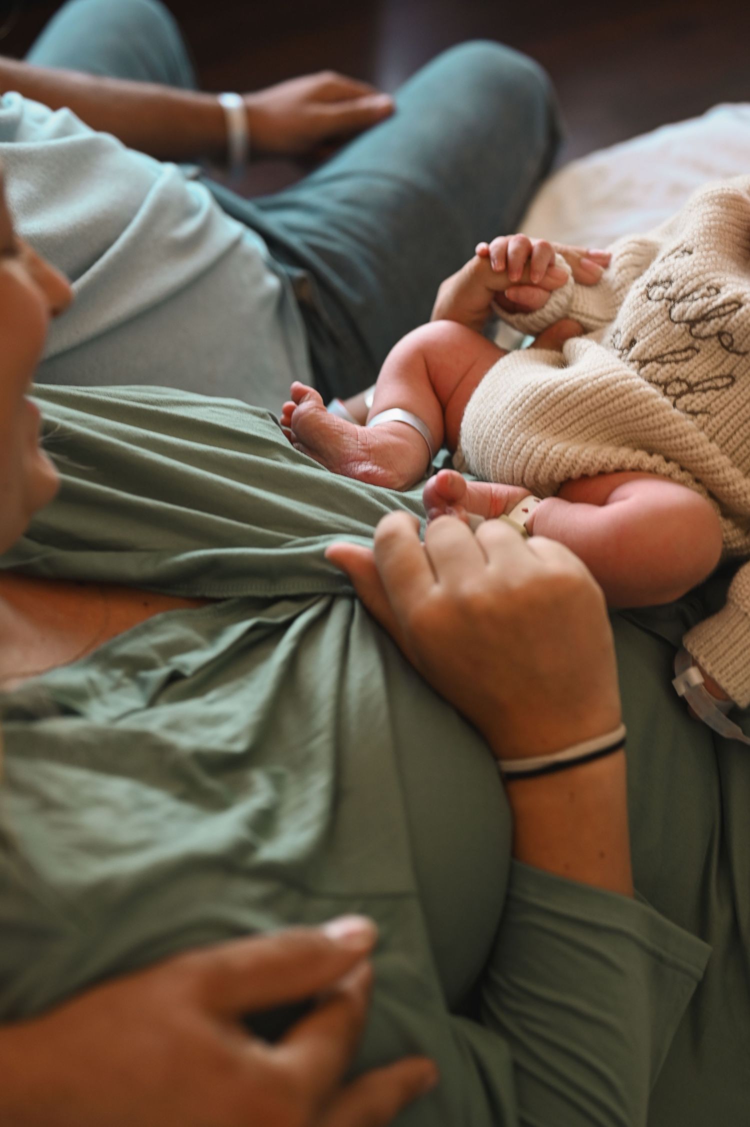 A newborn baby resting on a parent's lap while two adults gently hold the infant's tiny hands and feet.