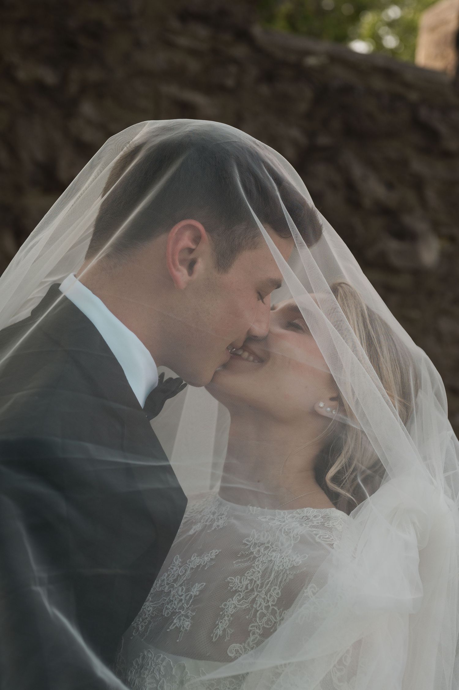Bride and groom sharing a romantic kiss under a flowing white wedding veil outdoors.