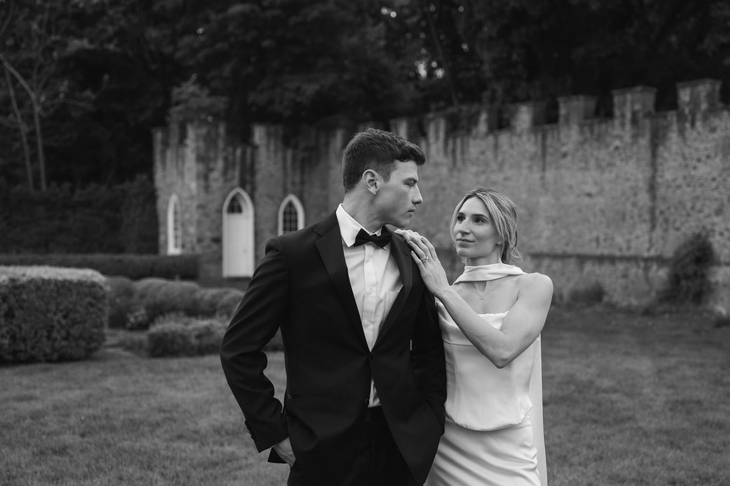 Black and white wedding photo of couple in formal attire posing outdoors near stone wall.