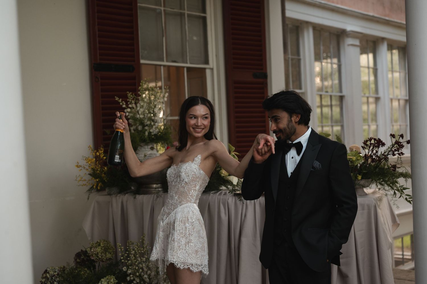 Bride in white lace mini dress holding champagne bottle with groom in black tuxedo at elegant wedding reception.
