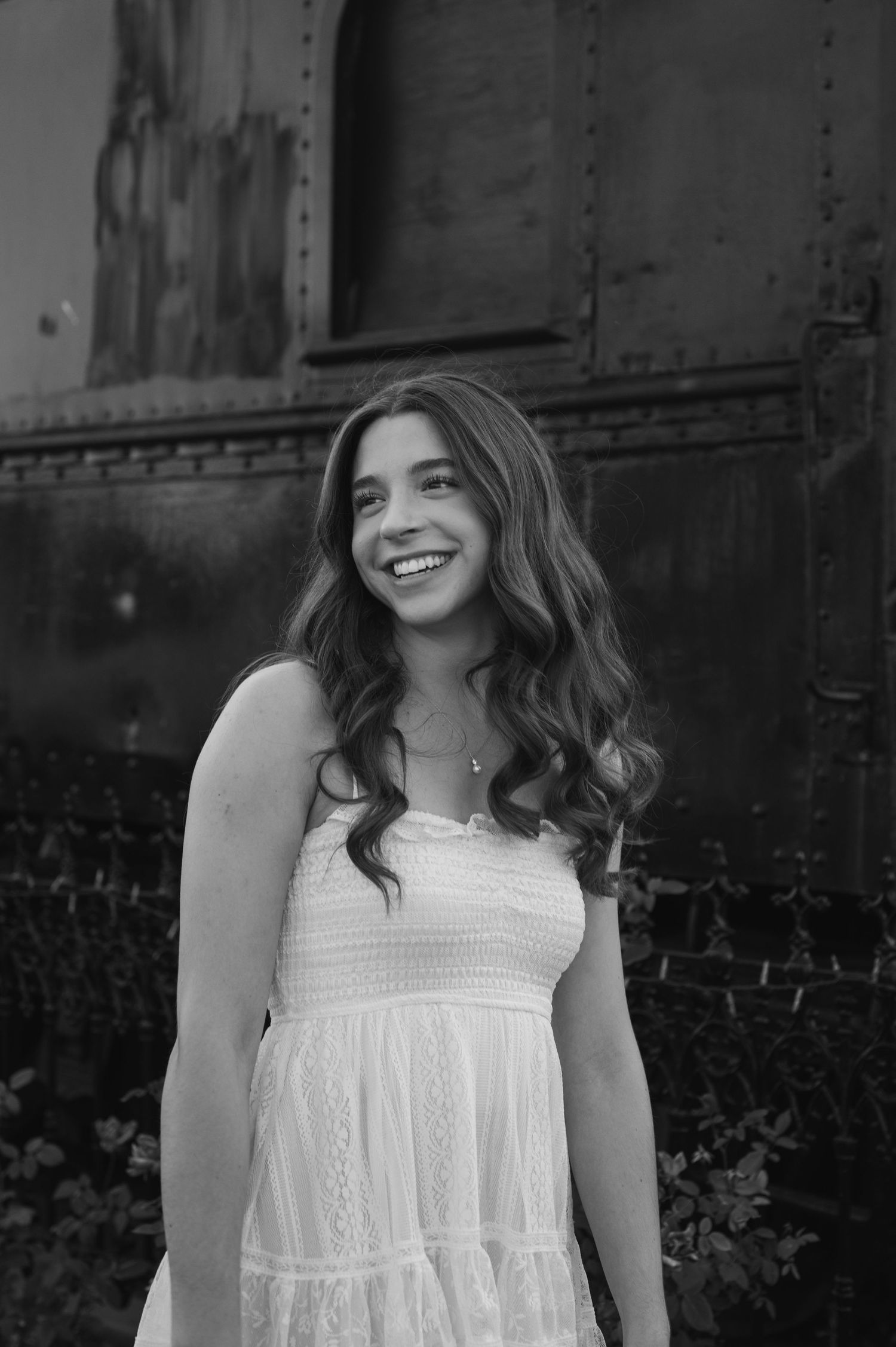 A young woman in a white sundress smiles joyfully outdoors in a black and white portrait photo.