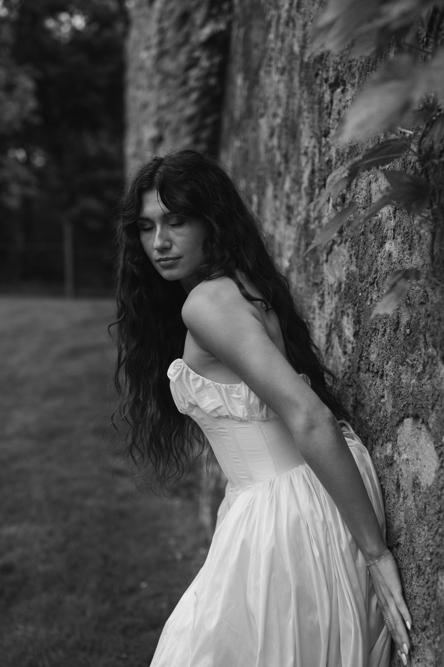 Black and white portrait of a young woman in a white dress leaning against a stone wall with long dark hair.