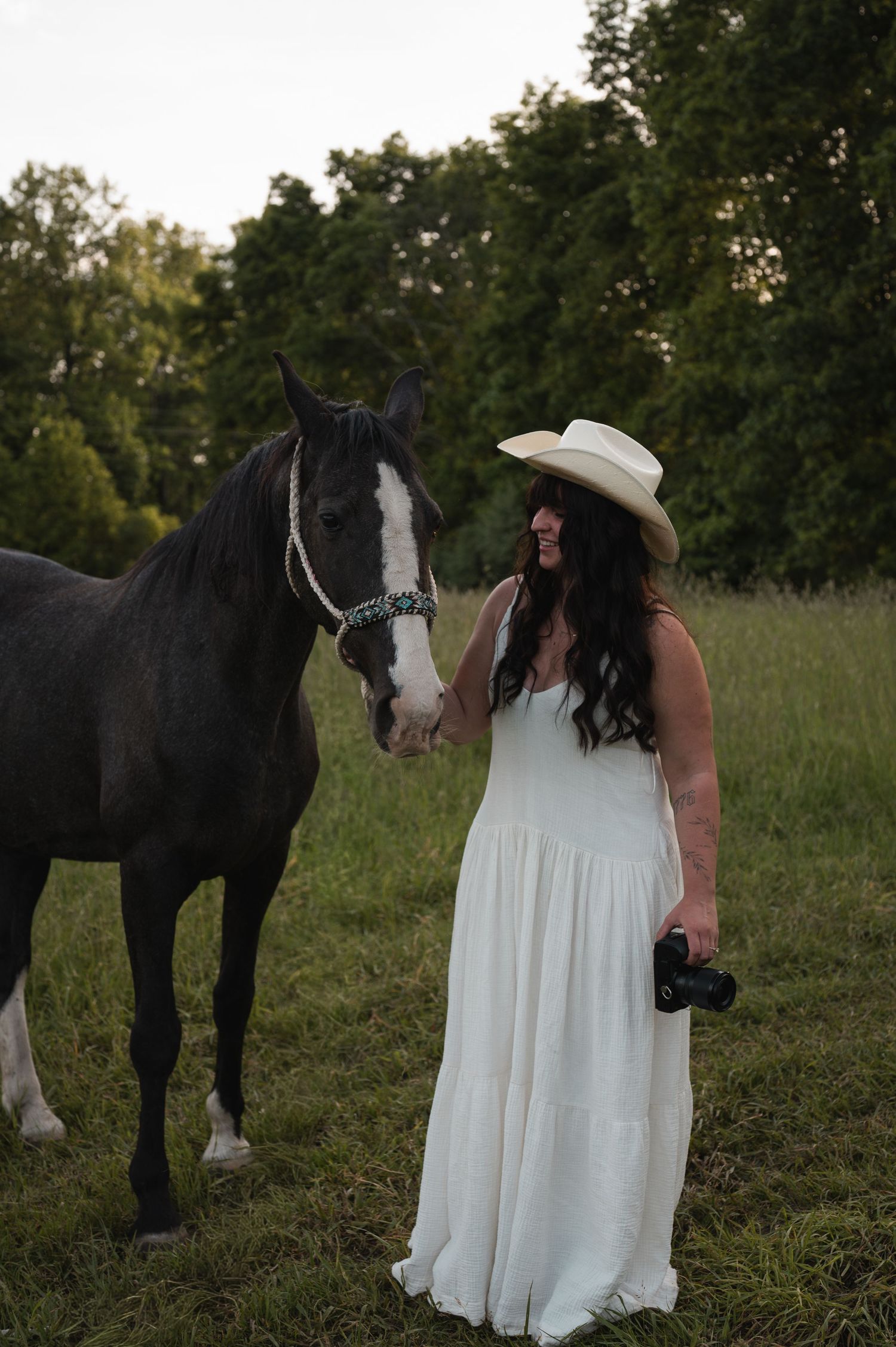Woman in white dress and cowboy hat stands beside a black horse in a green field at dusk, holding a camera.