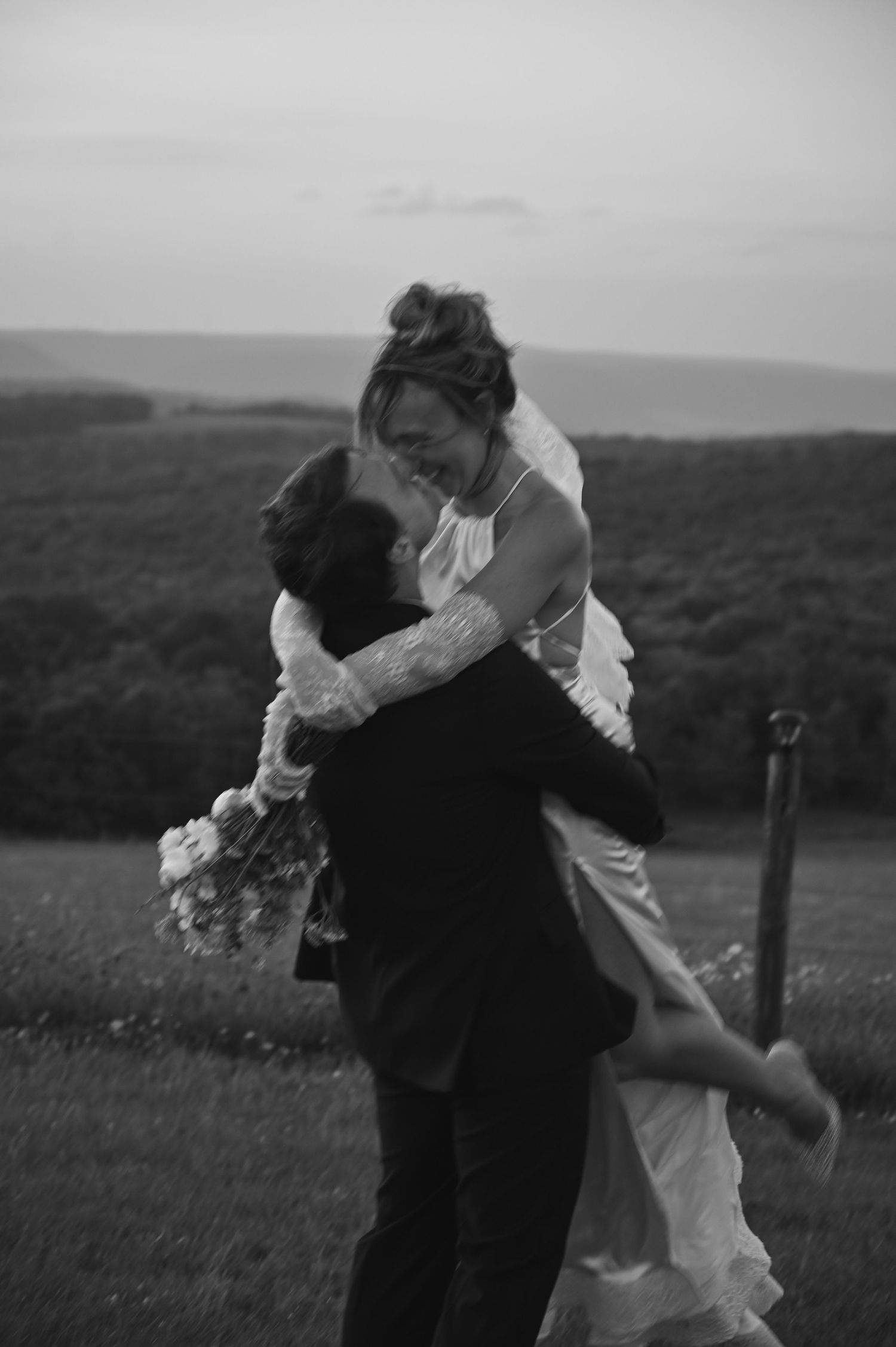 Black and white photo of a groom lifting his bride in a field, she laughs holding a bouquet on their wedding day.