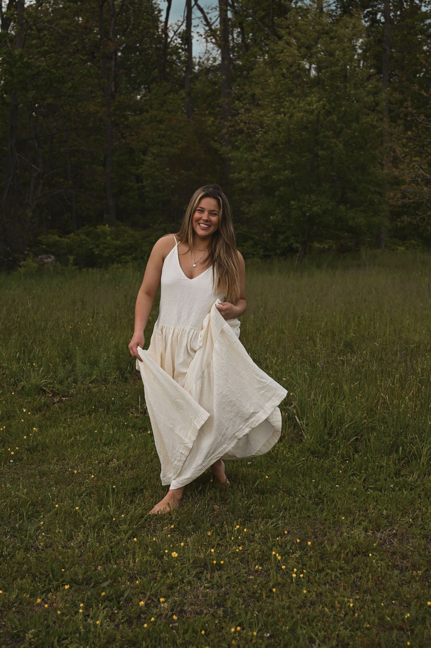 A smiling woman in a flowing cream maxi dress twirls barefoot in a lush green meadow surrounded by trees.