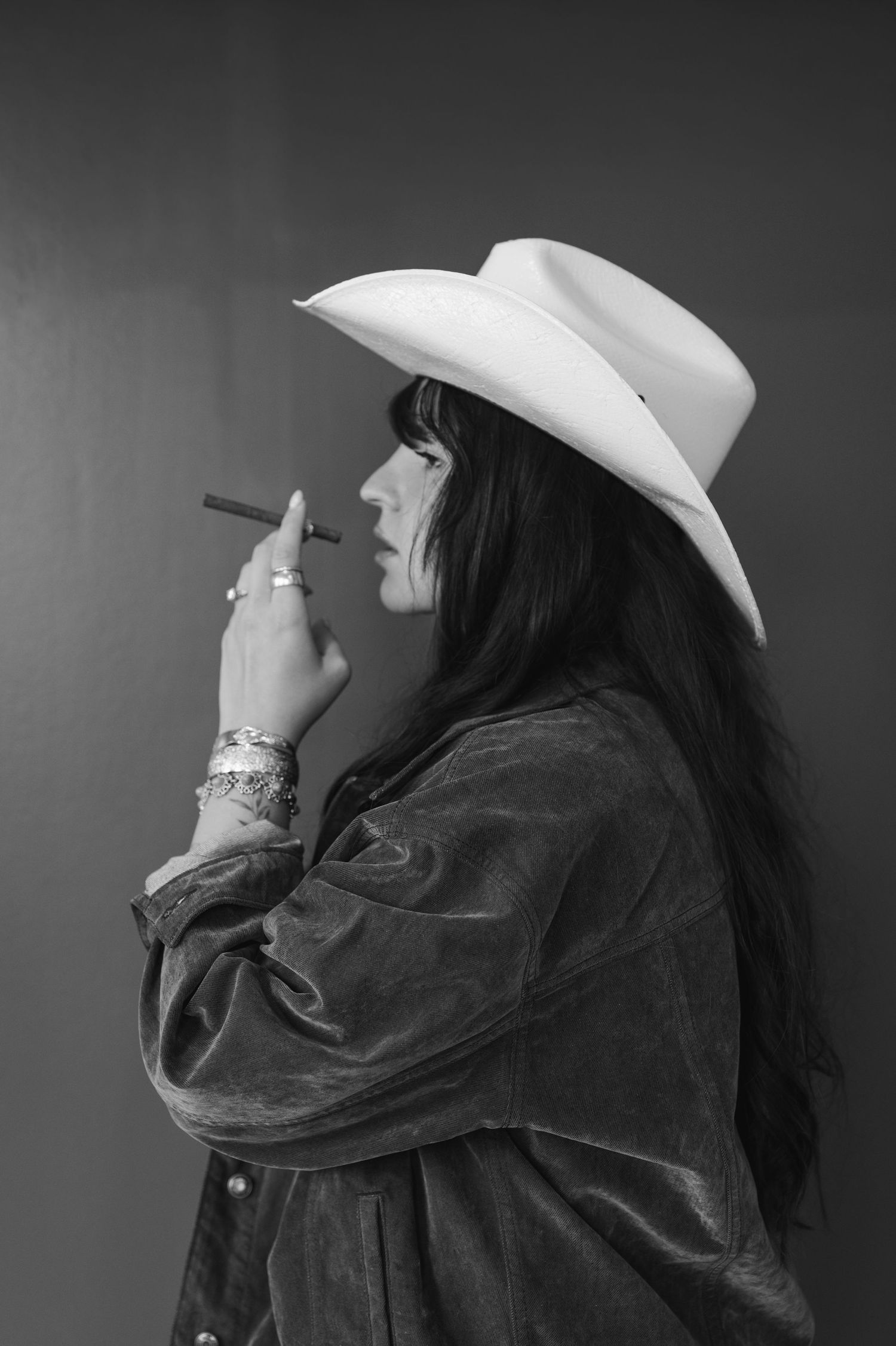 Black and white photo of a woman in a white cowboy hat and suede jacket smoking a cigar, viewed from the side.