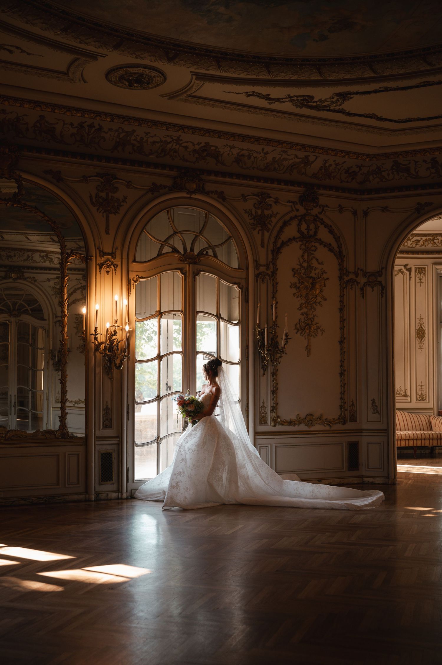 Bride in white gown with long train stands by ornate arched doors in an elegant baroque ballroom with golden decor.