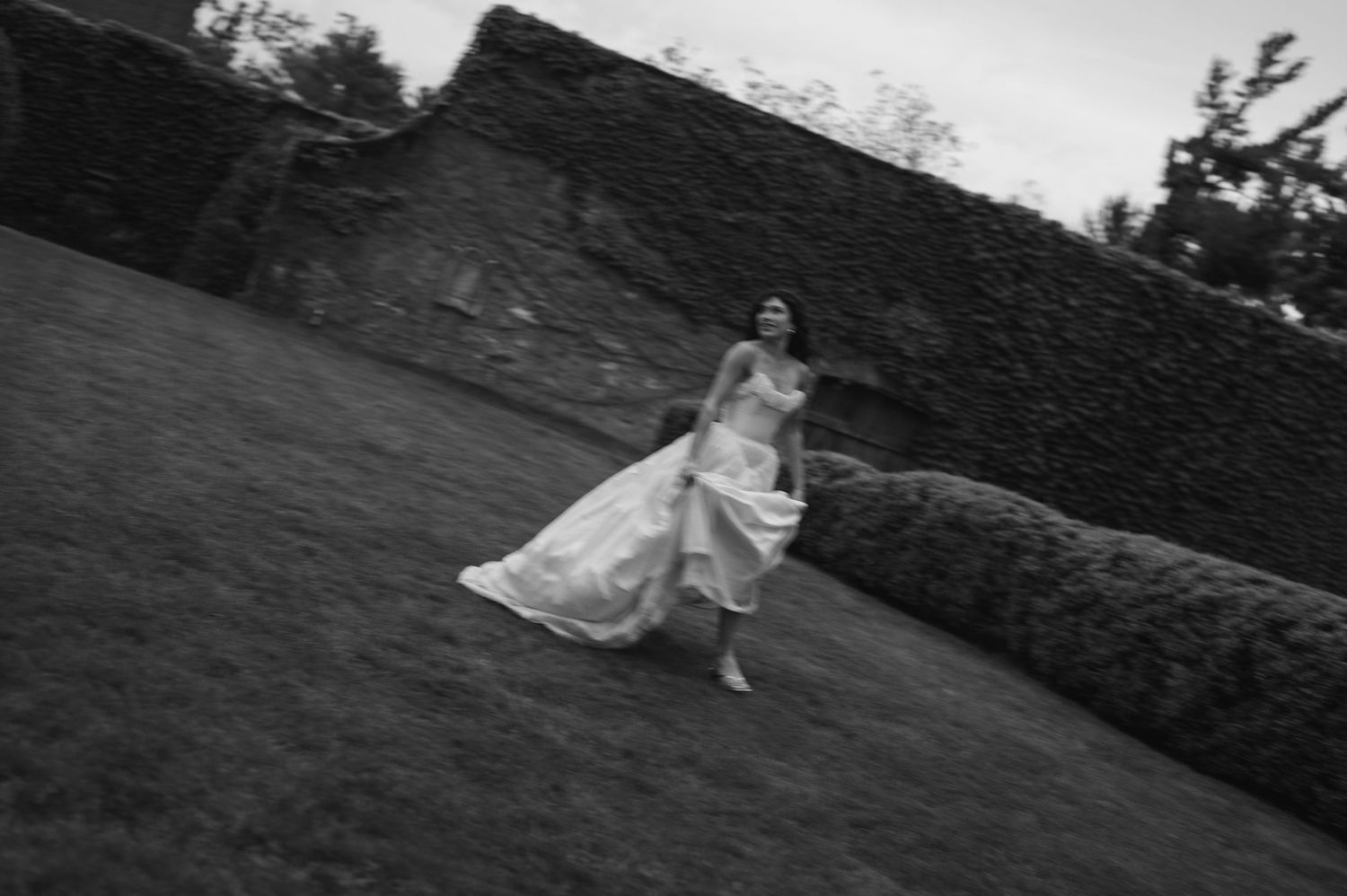 Black and white photo of a bride in a flowing gown running across a lush garden lawn near stone walls.