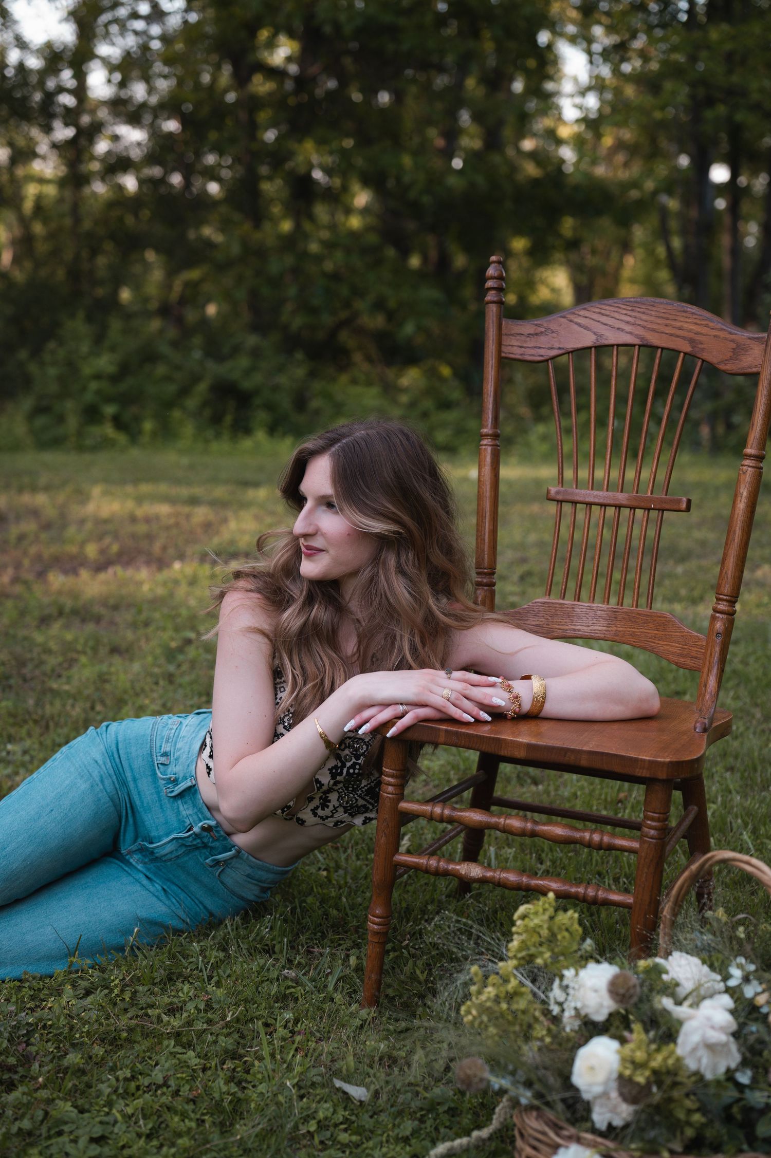 Woman in jeans resting on grass beside a wooden chair outdoors with flowers nearby.
