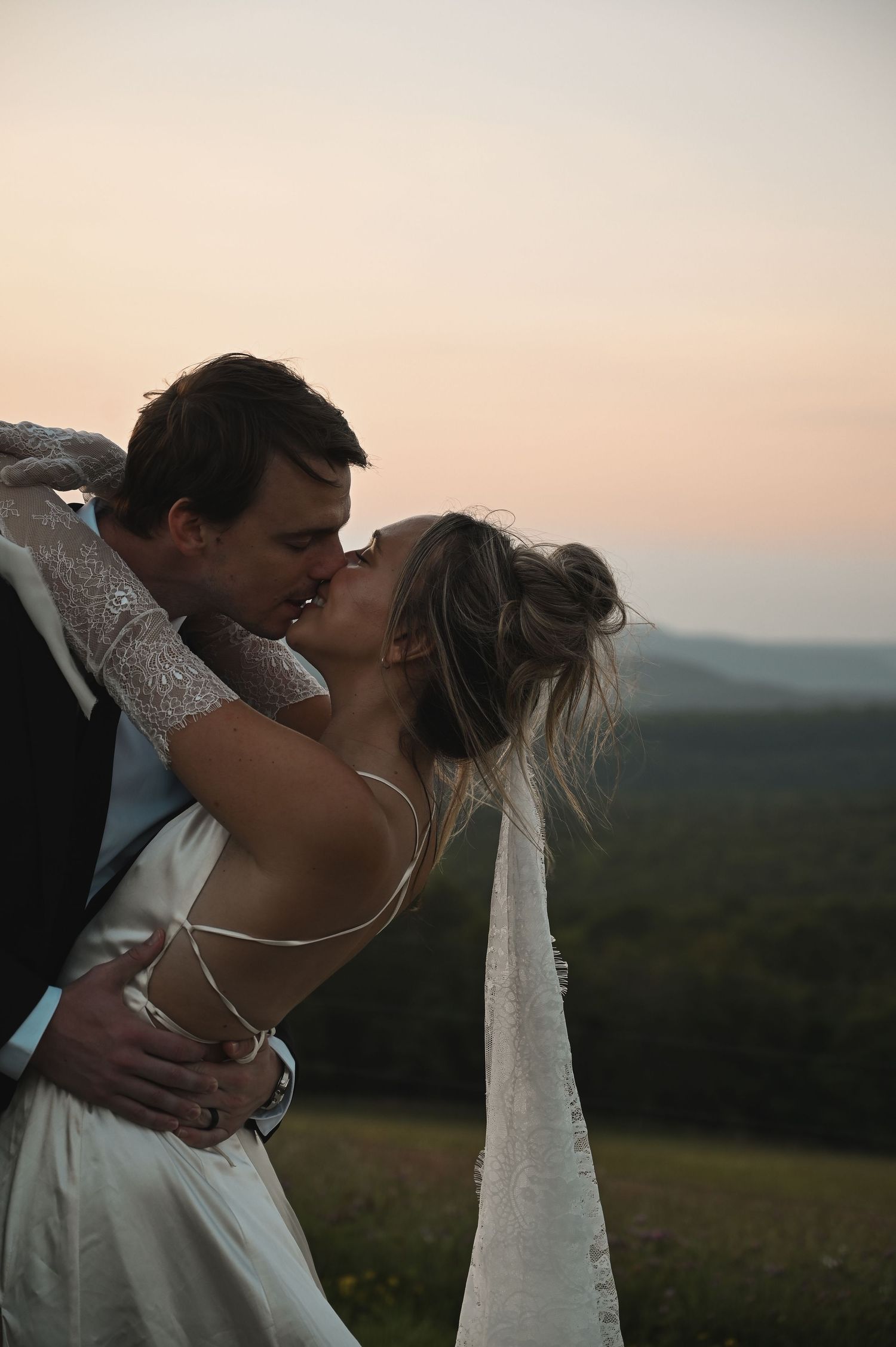 A romantic sunset moment captured with a couple in wedding attire embracing on a hilltop with mountains in the background.